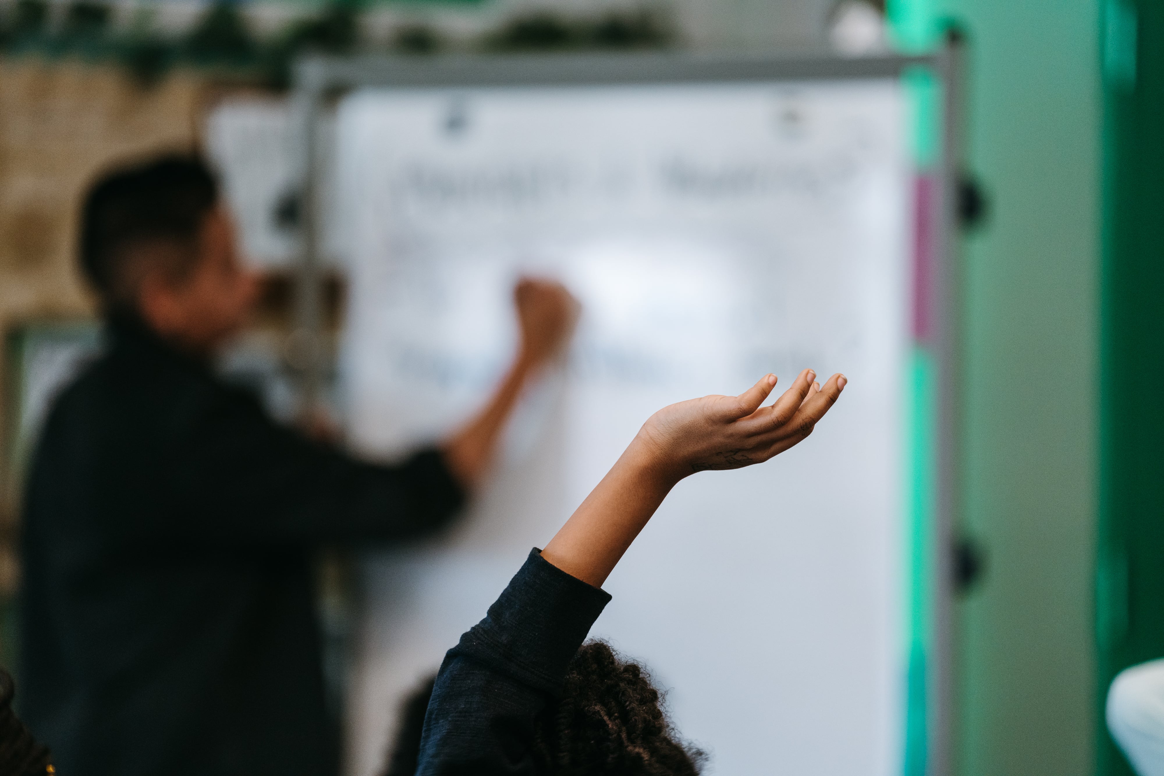 In the foreground, a student raises their hand. Their face is not visible. In the background, a teacher writes on the whiteboard in a middle school classroom.