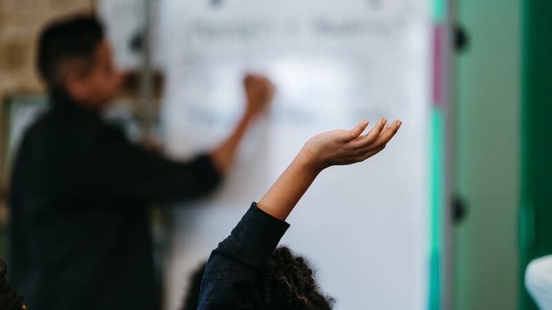 In the foreground, a student raises their hand. Their face is not visible. In the background, a teacher writes on the whiteboard in a middle school classroom.