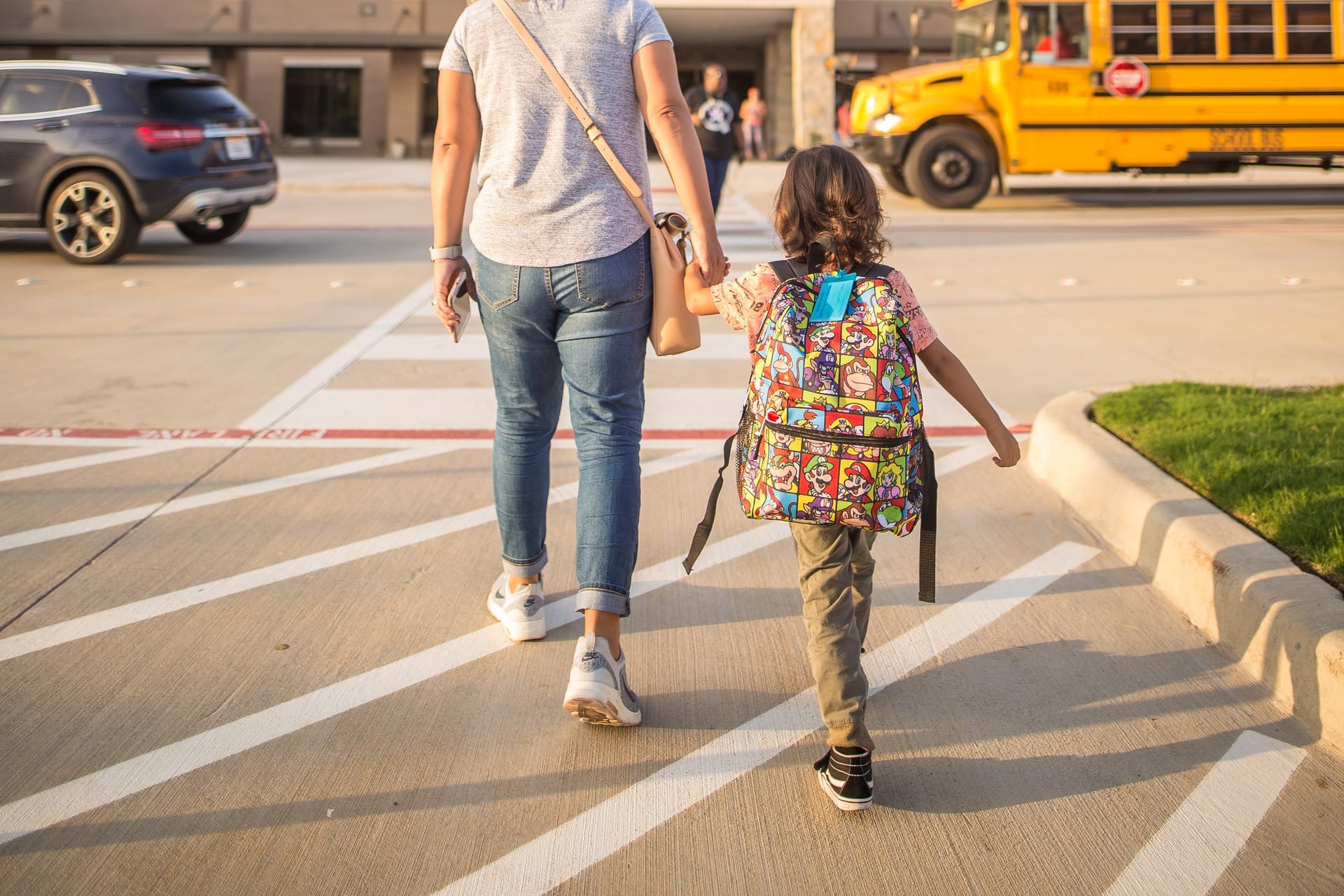 Mother and child holding hands walking toward school entrance. A school bus in the background.