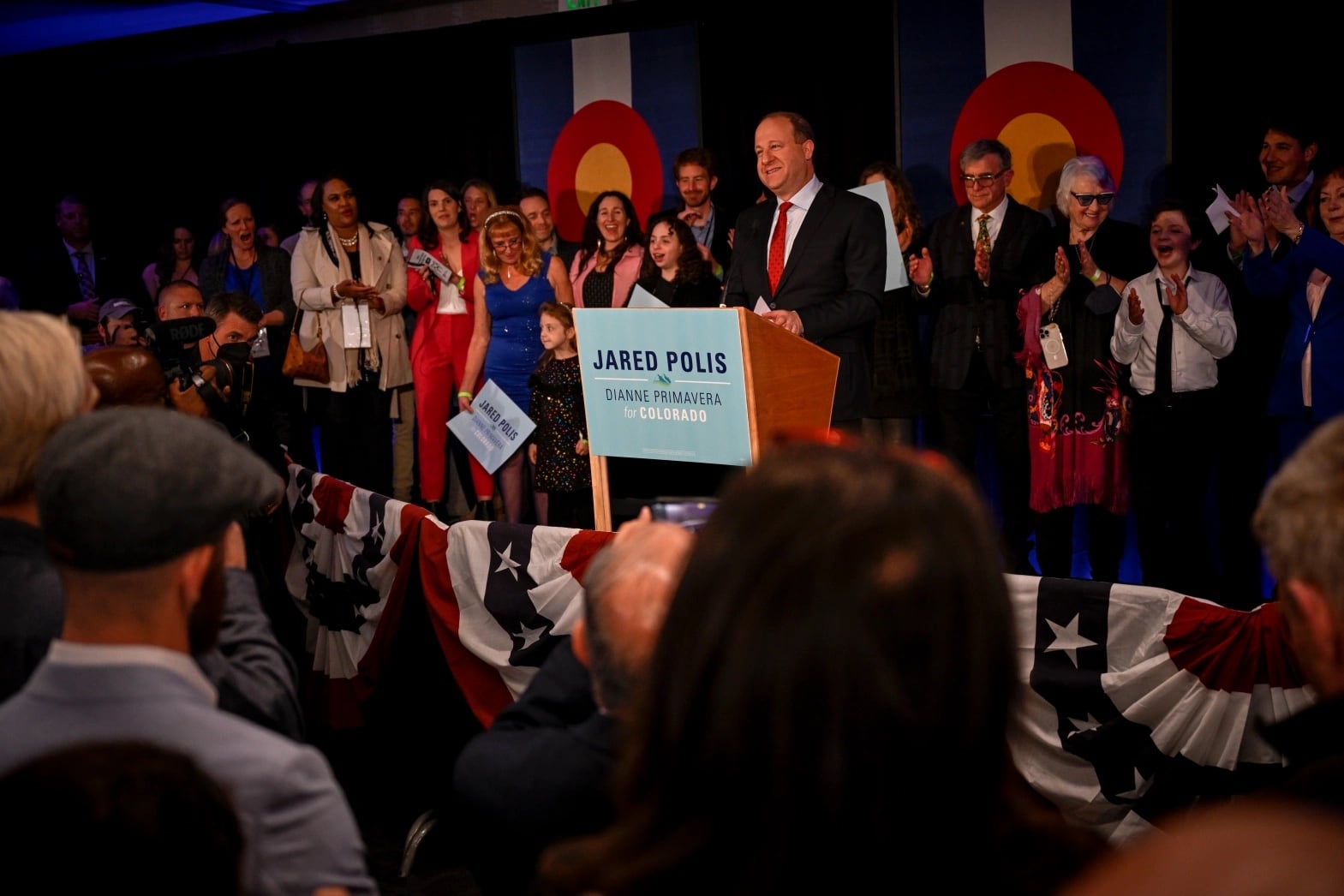 Colorado Gov. Jared Polis speaks from a podium with a large crowd behind him. Colorado flags hang from the wall behind them. 