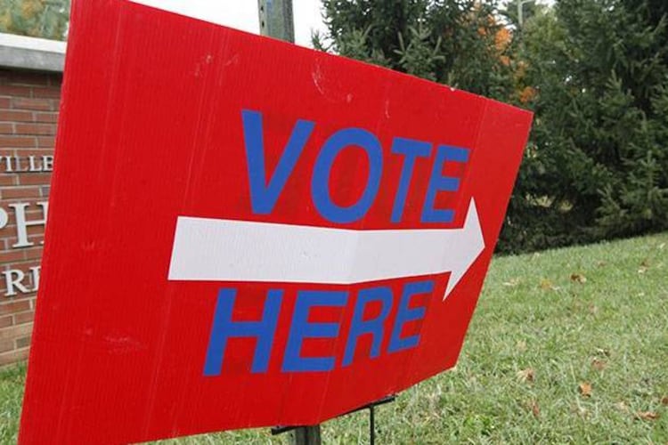 A red sign with blue “Vote Here” letters and a white arrow. 