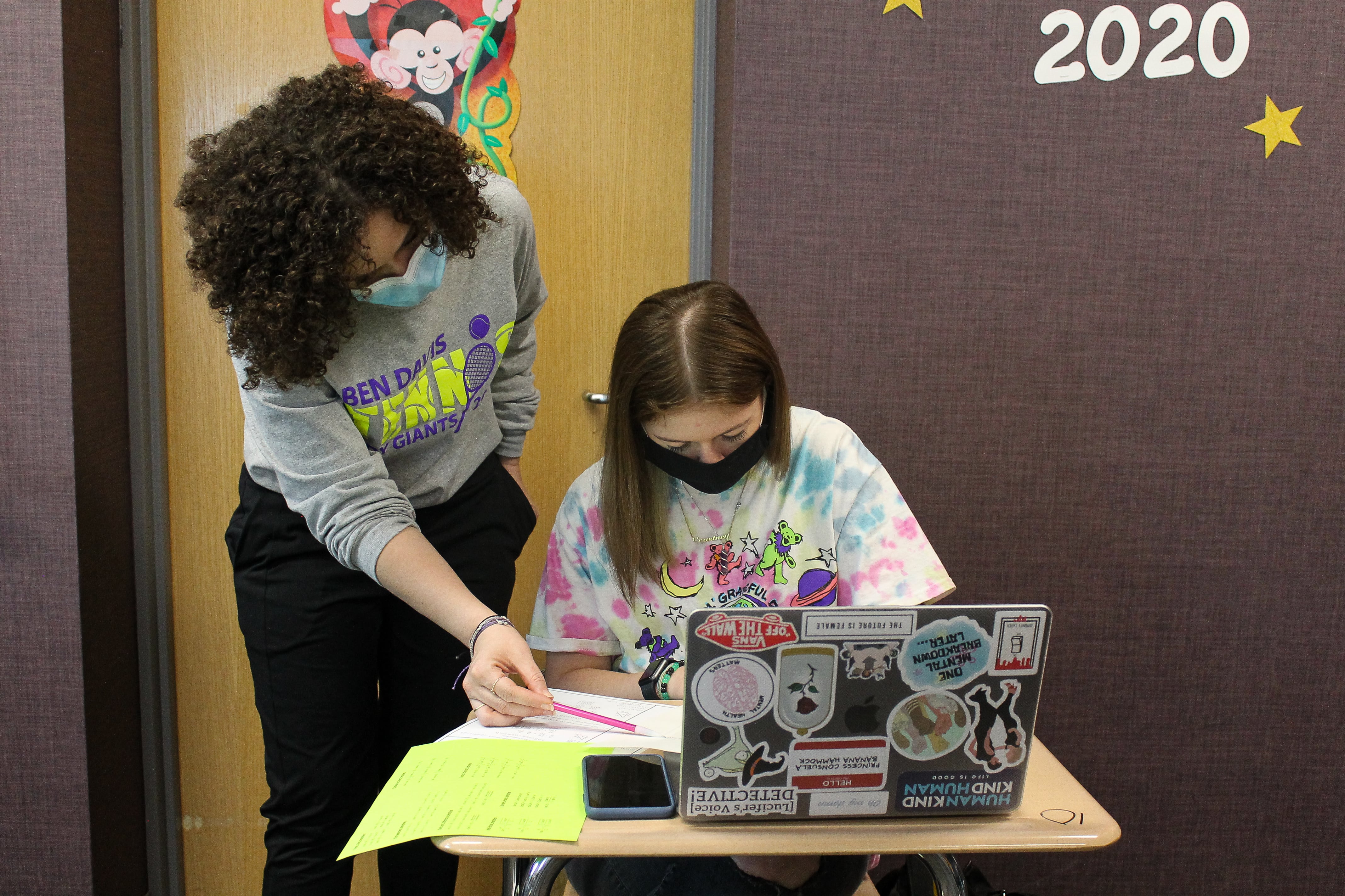A masked teacher helps a masked student at a desk.