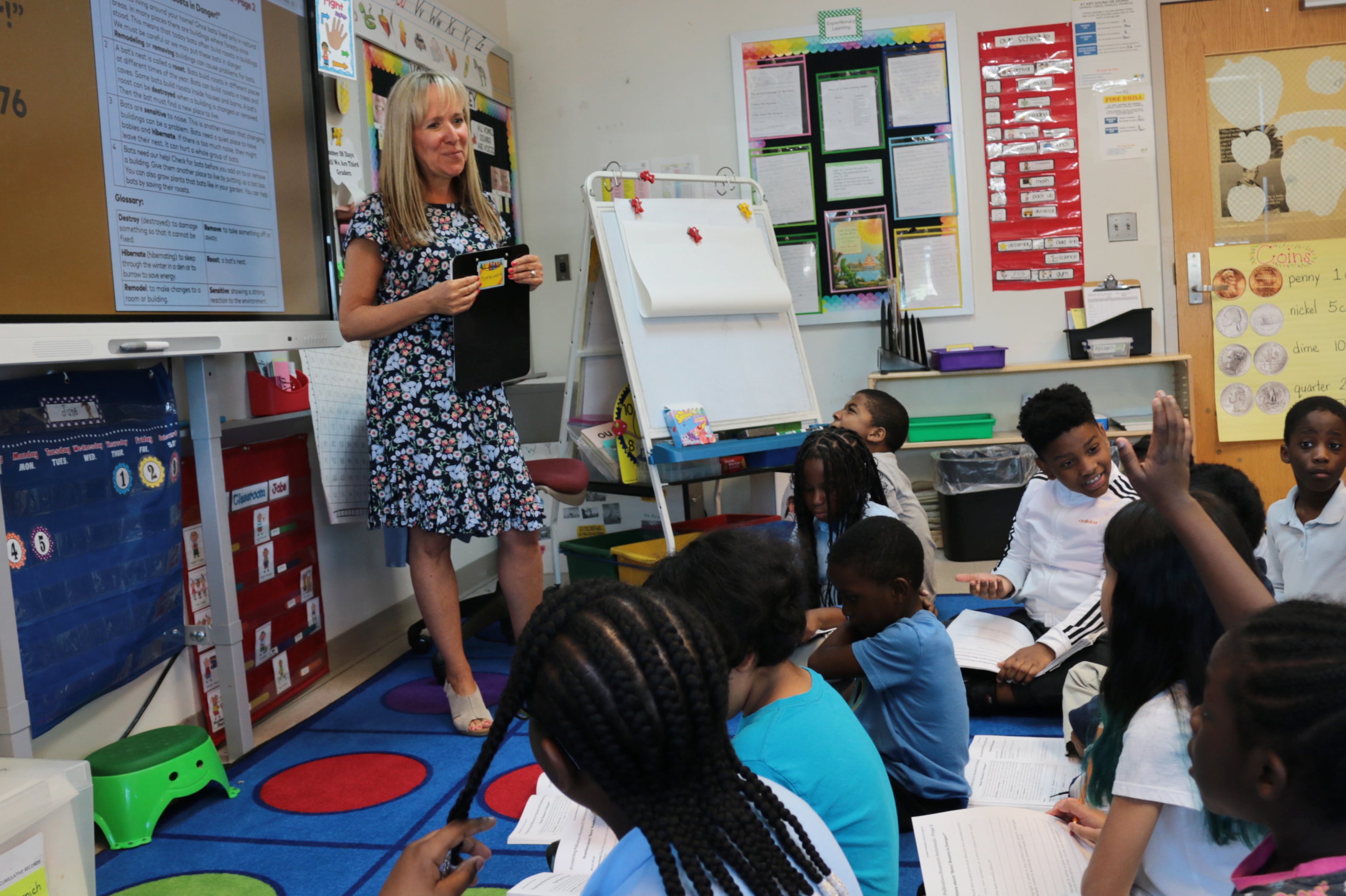 A blonde woman in a blue patterned dress stands in front of a classroom of students.