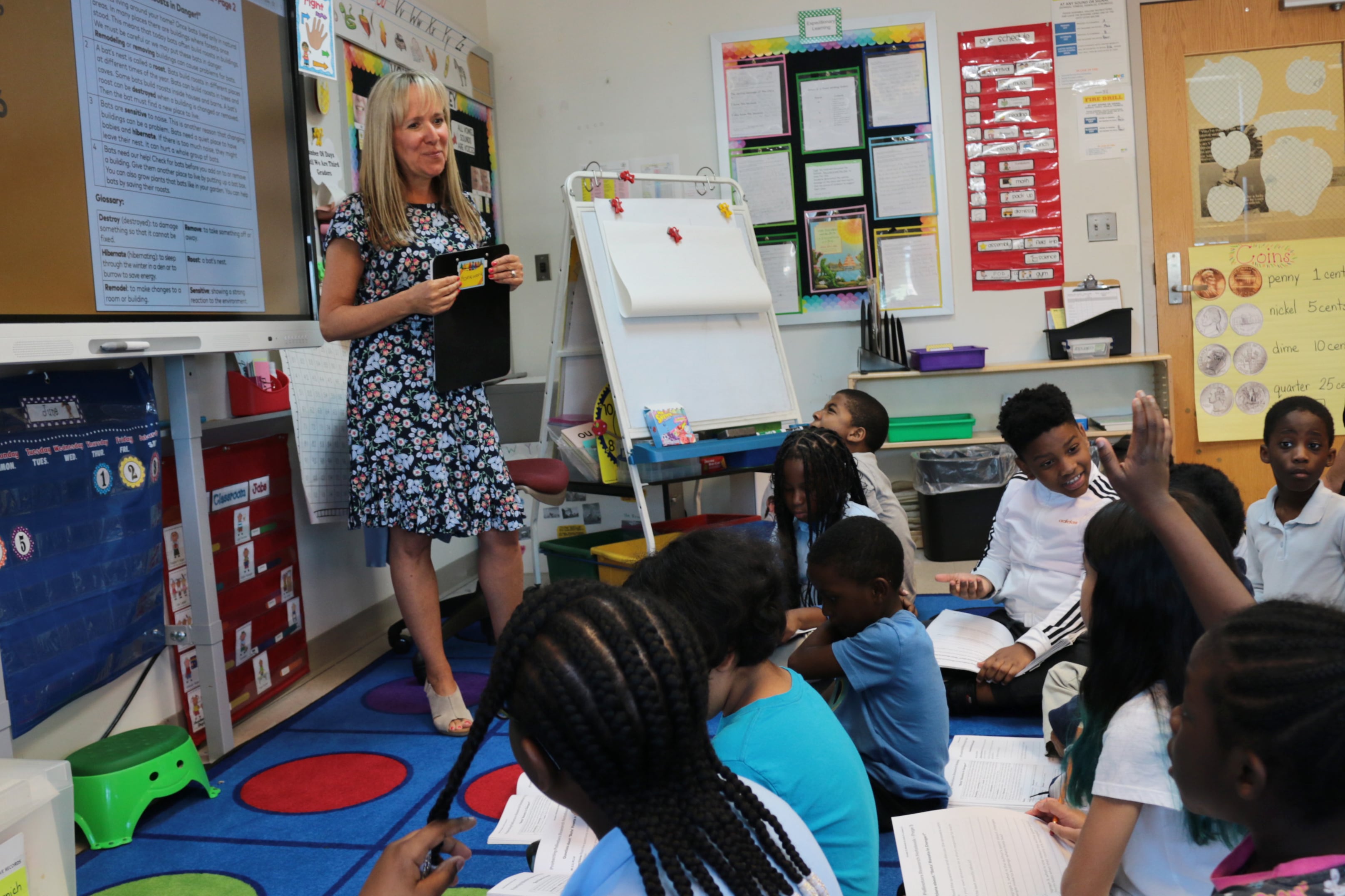 A blonde woman in a blue patterned dress stands in front of a classroom of students.