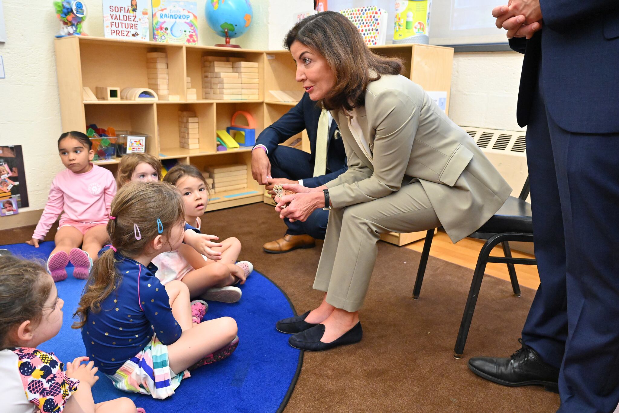 A woman in a tan suit sits on a chair and leans toward a group of young children on a carpet on the floor.