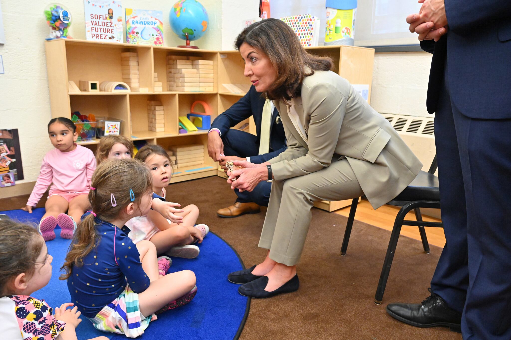 A woman in a tan suit sits on a chair and leans toward a group of young children on a carpet on the floor.