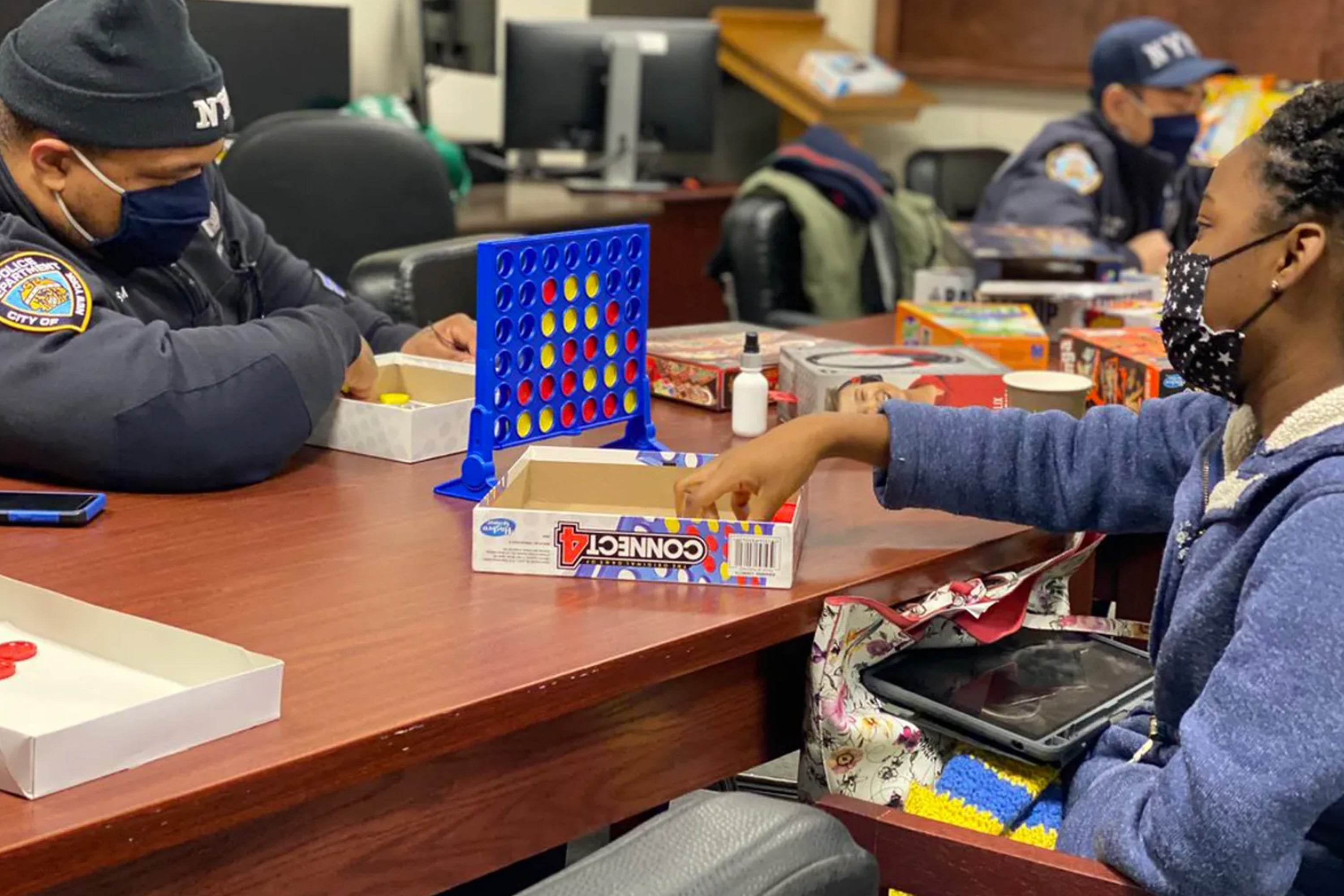 A police officer sits at a wood table playing Connect Four with a girl. 