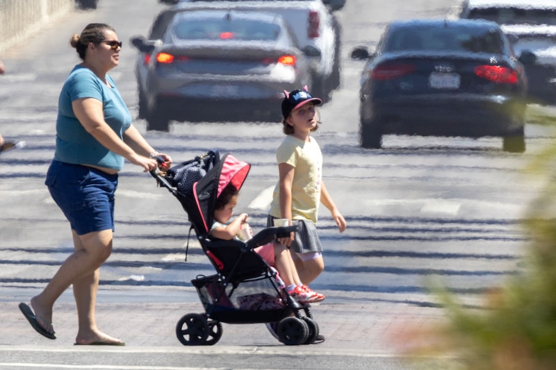 An adult woman pushes a young child in a stroller and walks alongside a young child across a busy intersection with a line of cars driving away in the background.