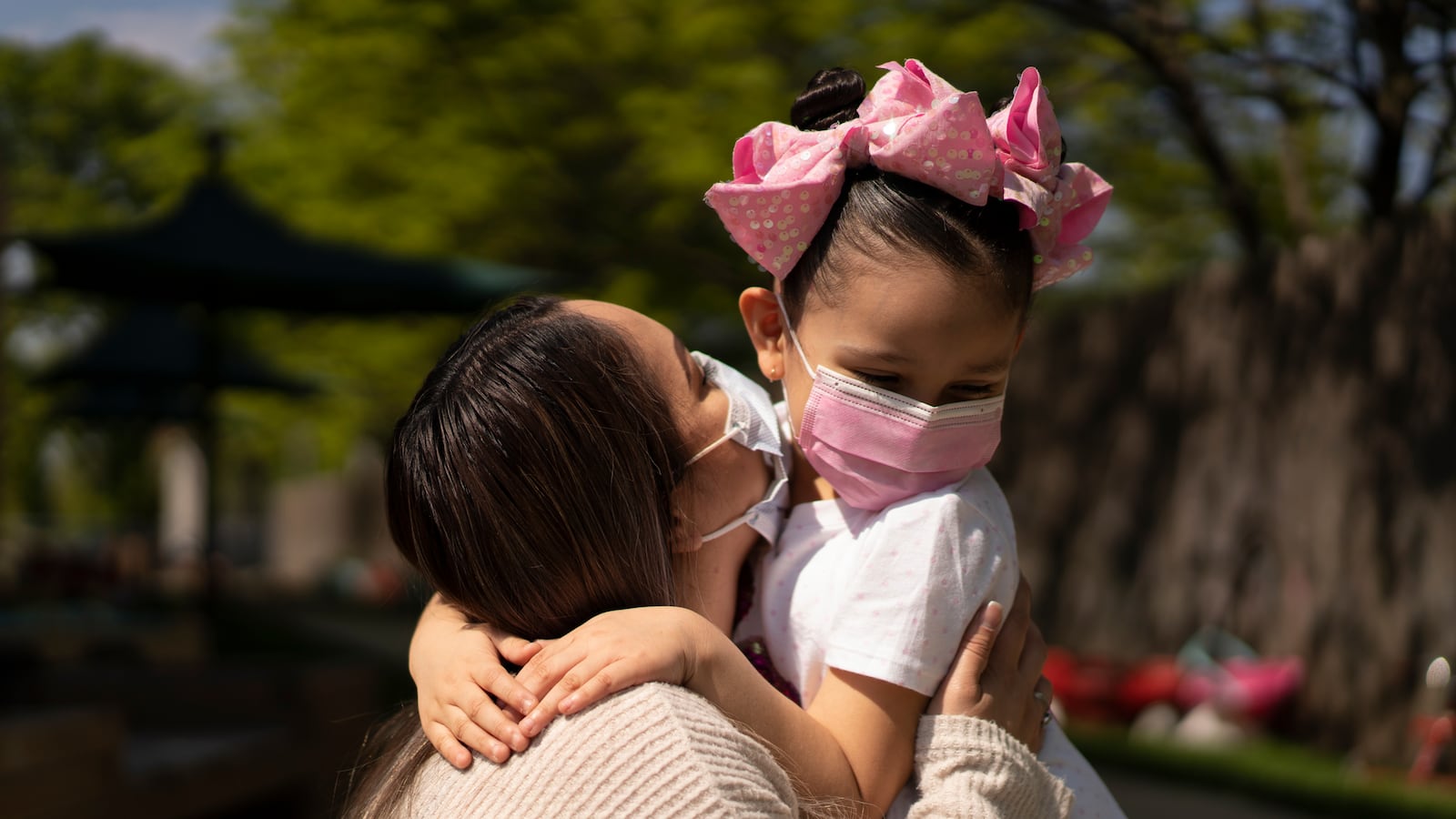 A mother wearing a tan shirt and a protective mask holds her daughter, who is wearing a white shirt, pink mask and pink polka-dot bow. The background is a swirl of green trees.