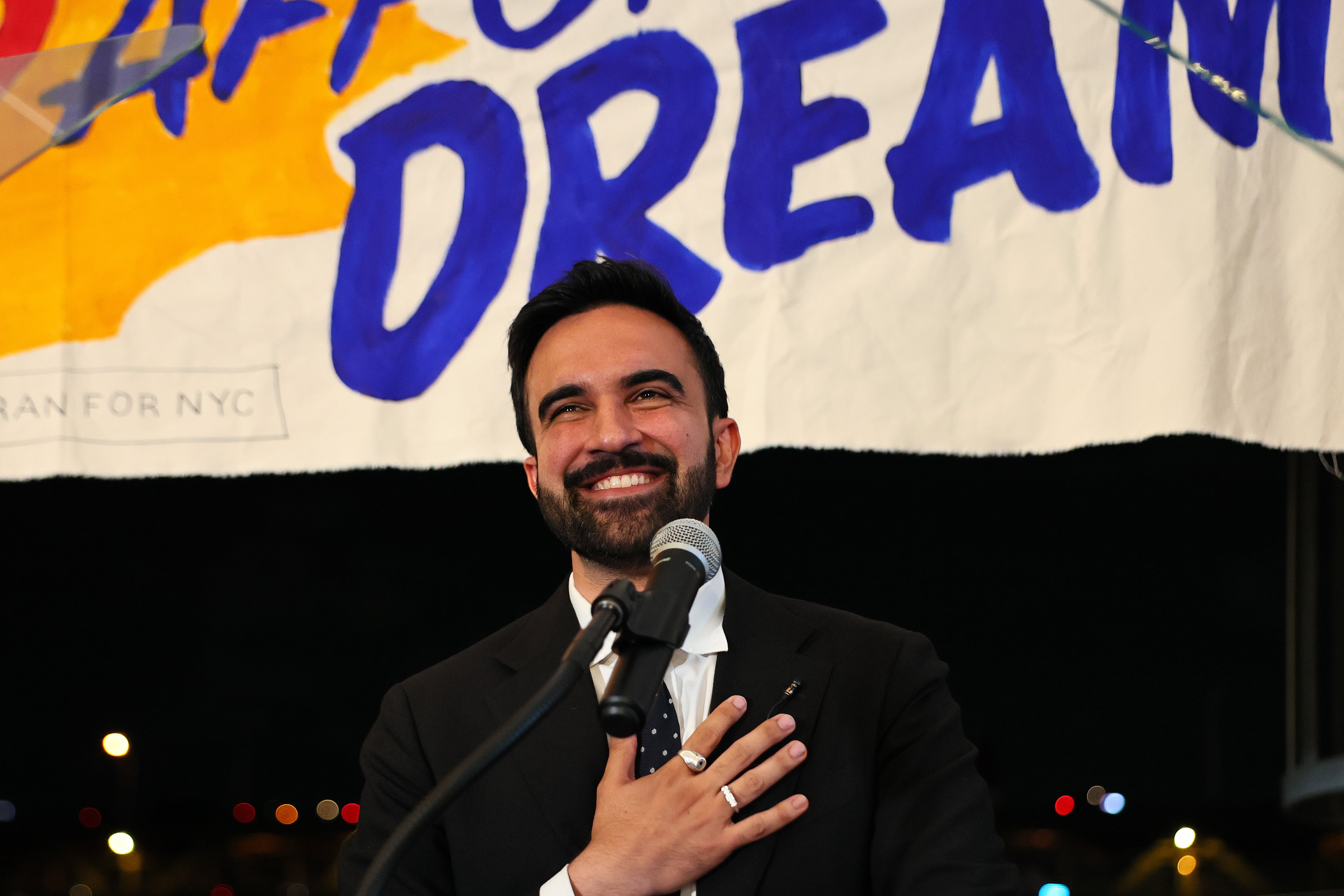 A man in a dark suit holds his hand to his chest in front of a banner.