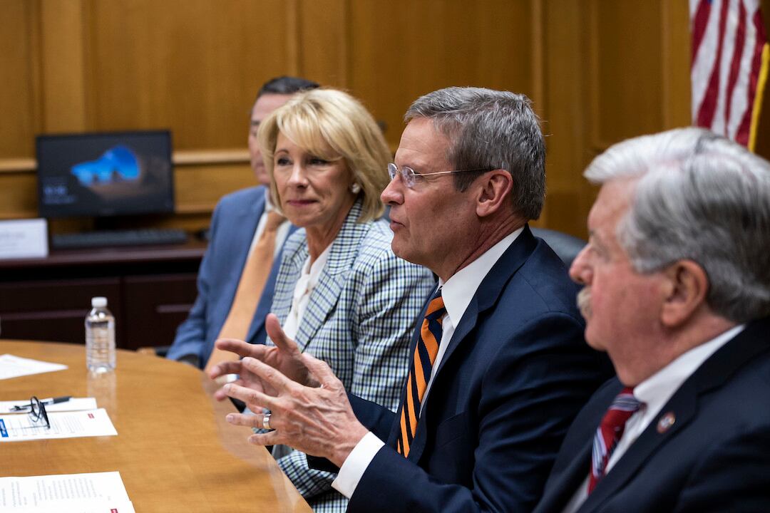 a man in a suit and orange striped tie speaks and gestures at a table while Betsy DeVos wearing a checked suit looks on in the background