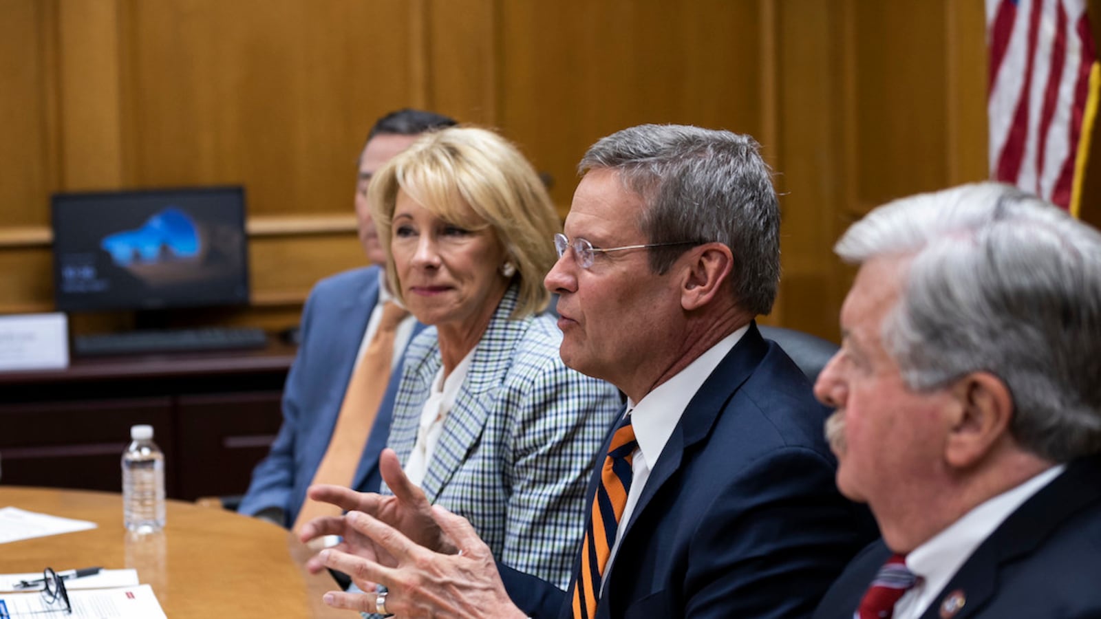 a man in a suit and orange striped tie speaks and gestures at a table while Betsy DeVos wearing a checked suit looks on in the background