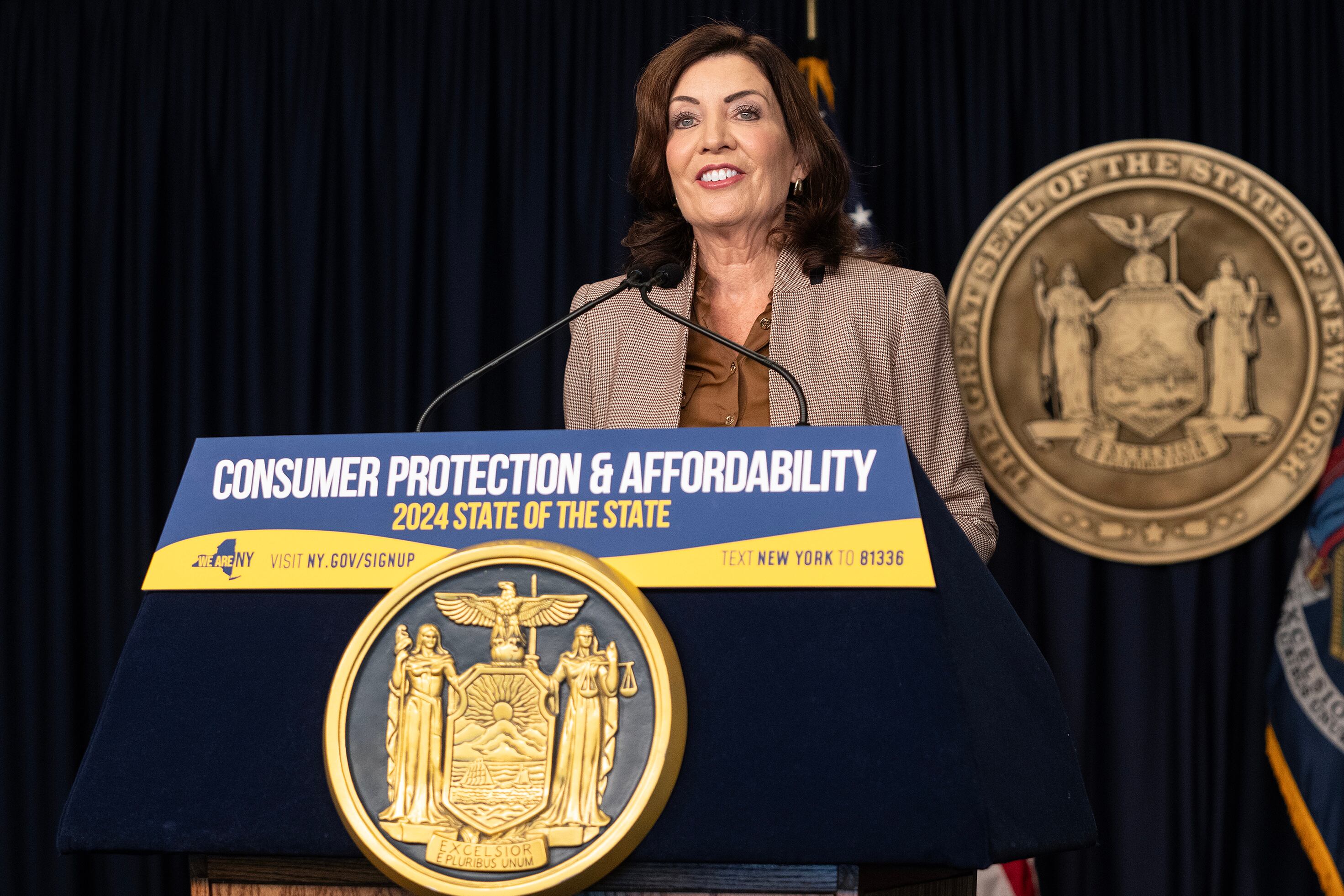 A woman with dark brown hair and wearing a light brown suit jacket stands behind a podium and speaks into a microphone. There is the New York State seal in the background and in the foreground.
