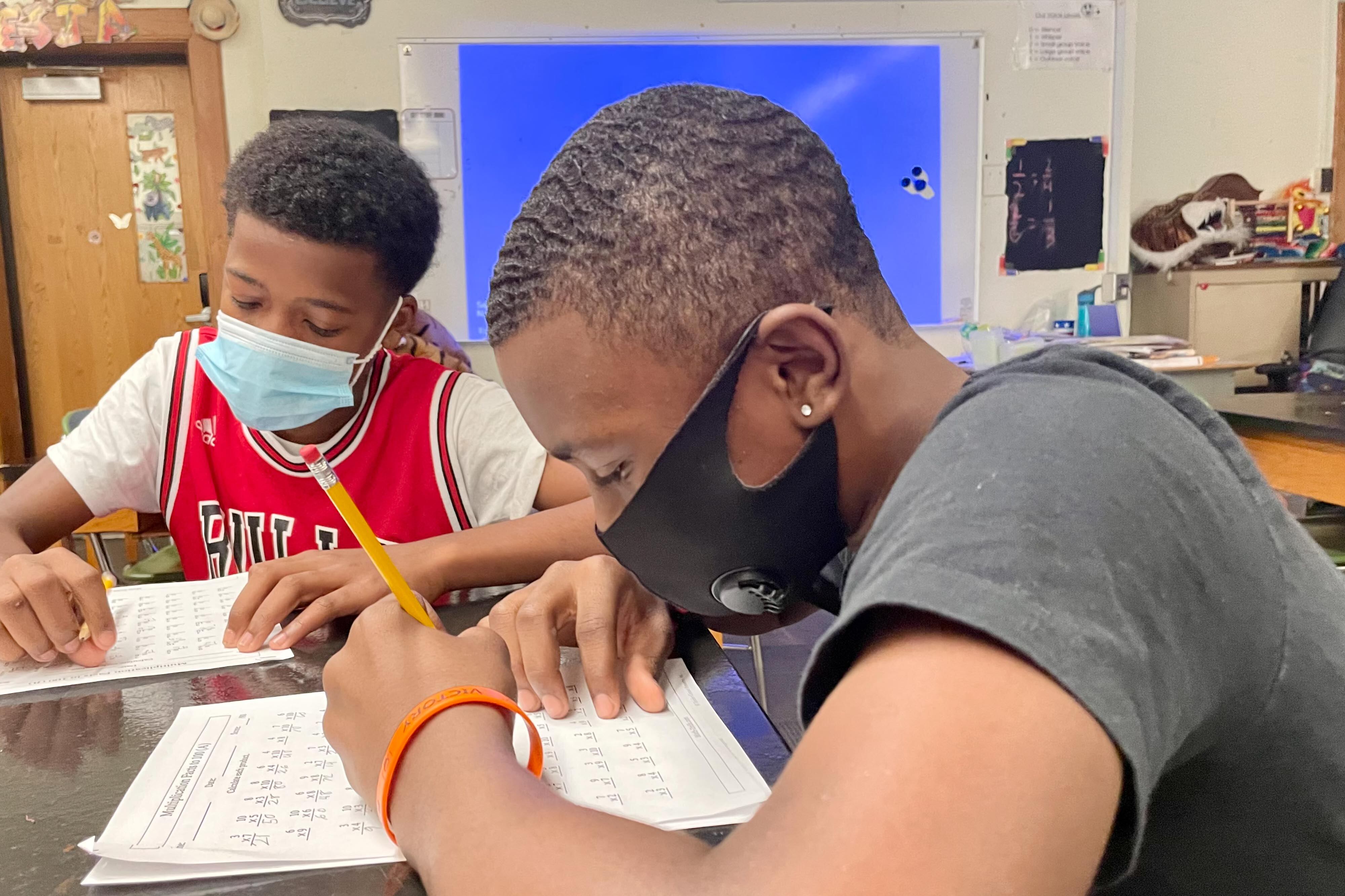 Two male middle school students work on mathematics worksheets at their desk. Both are wearing face masks.