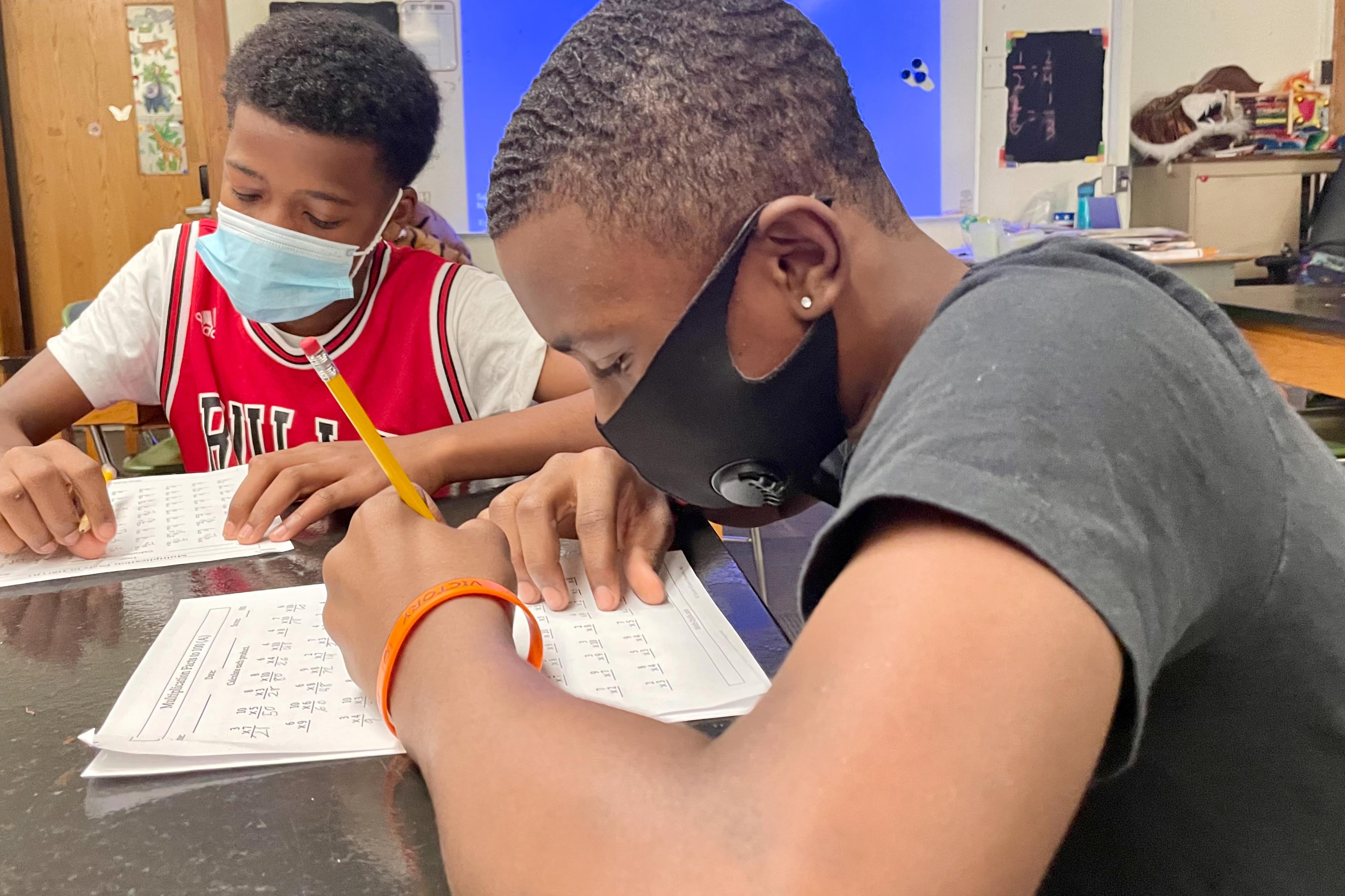 Two male middle school students work on mathematics worksheets at their desk. Both are wearing face masks.