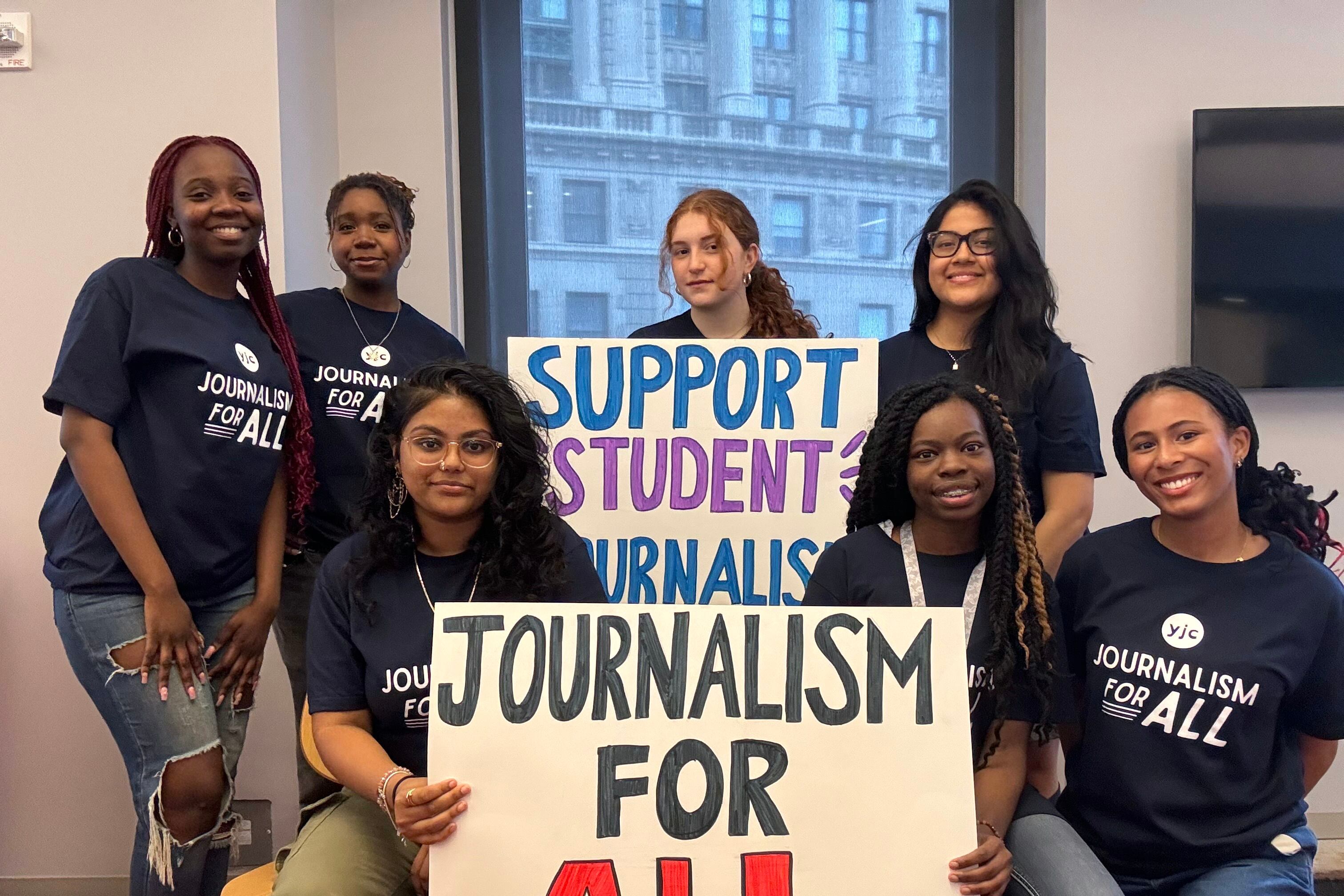 A group of young journalists wearing matching blue shirts pose for a photograph standing around two signs, one reads "Journalism for All," and Support Student Journalists."