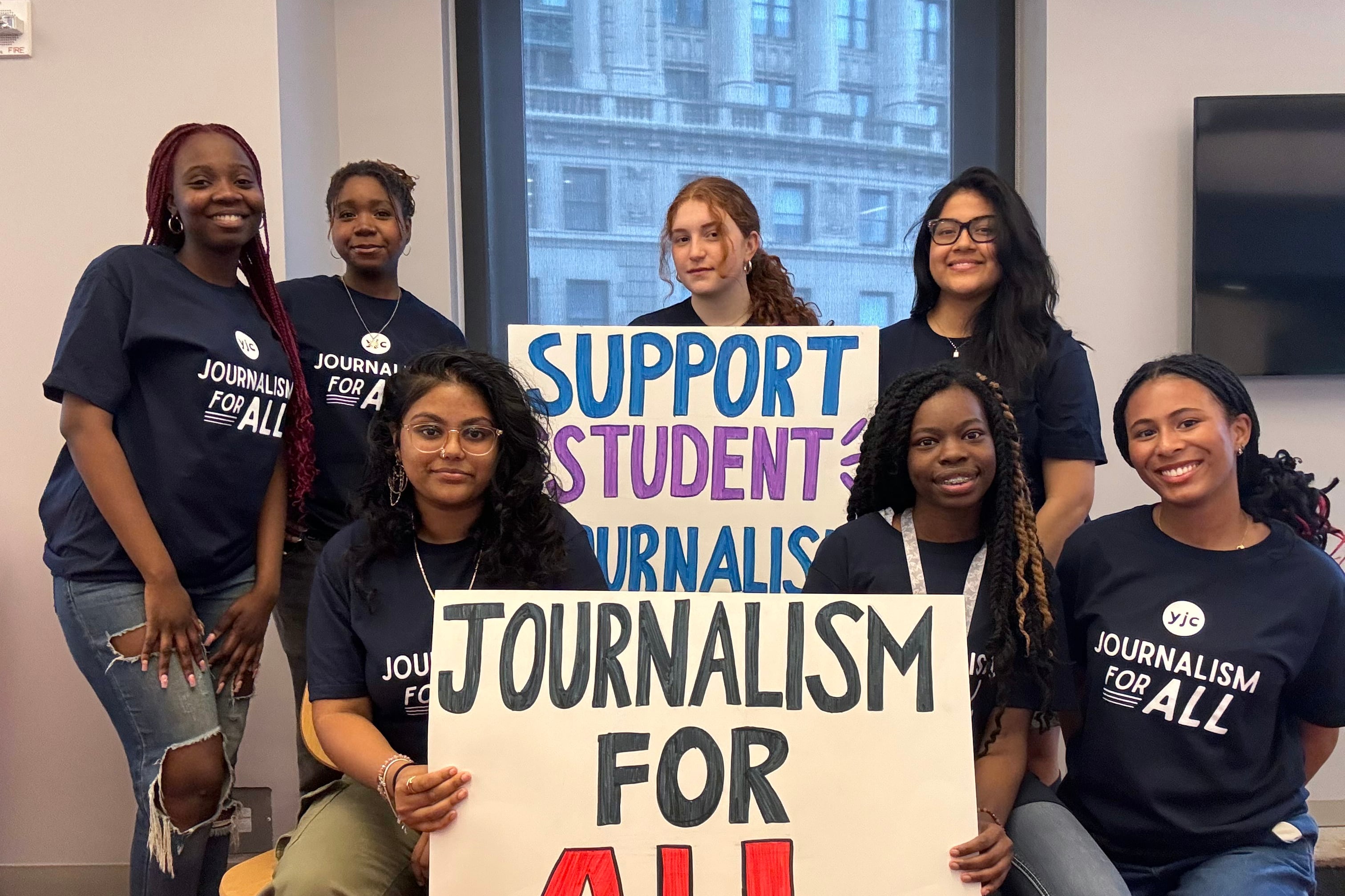 A group of young journalists wearing matching blue shirts pose for a photograph standing around two signs, one reads "Journalism for All," and Support Student Journalists."