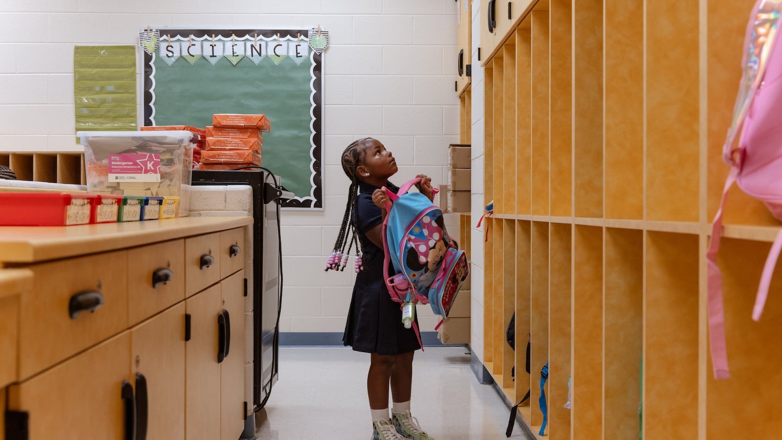 A young student with long braids stands between a desk and wooden cubbies and lifts her blue and pink backpack into her cubby. There is a white brick wall with a green board in the background.