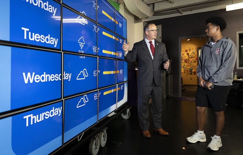 A photograph of an adult white man in a suit stands next to a Black student in a weather report room.