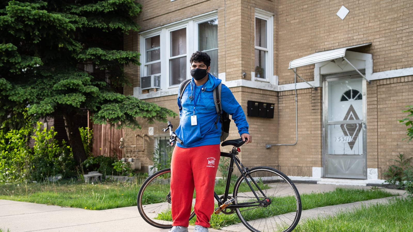 A young man, wearing a blue jacket, protective mask, and red track pants, poses for a portrait with his bike before riding to his high school.