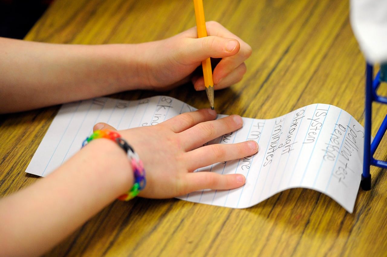 A close up of a young student's hands writing on a piece of paper on a wooden desk.