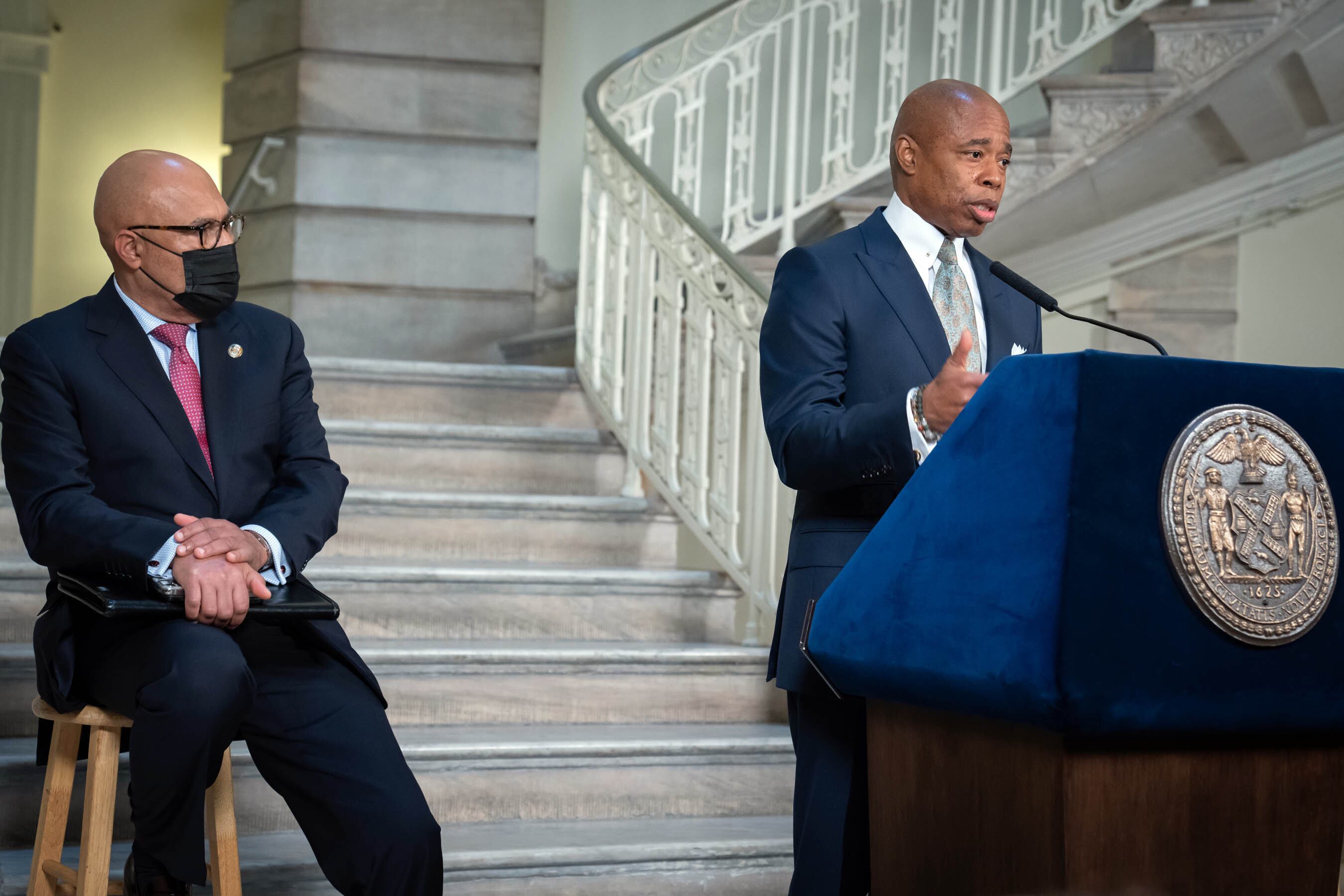 NYC Mayor Eric Adams speaks at a podium during a press conference, in front of a large staircase, as a man wearing a mask and glasses sits on a stool behind him.