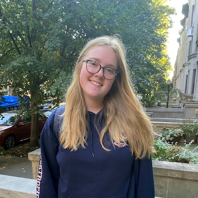 A high school student with long blonde hair and wearing glasses poses for a portrait outside with green trees and brick buildings in the background.