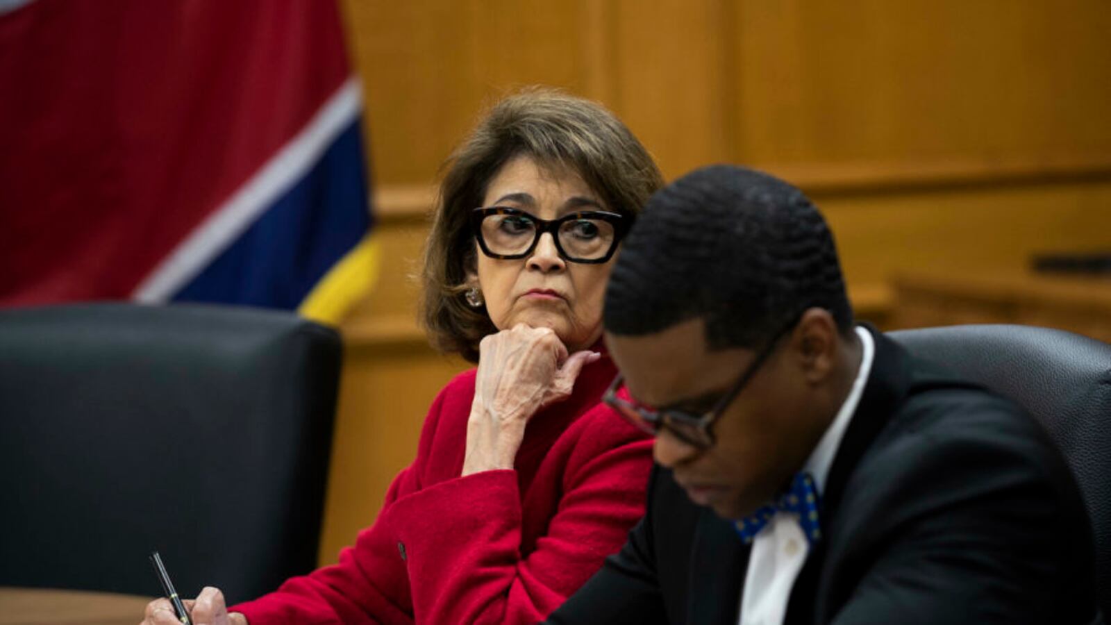 Two people sit at a wooden desk holding pens and writing on paper. A wood wall and part of a flag in the background.
