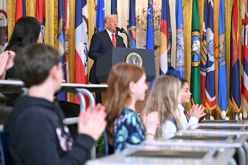 A white man in a dark suit speaks from a podium with young children sitting at desks in the foreground and a wall of colorful flags in the background.