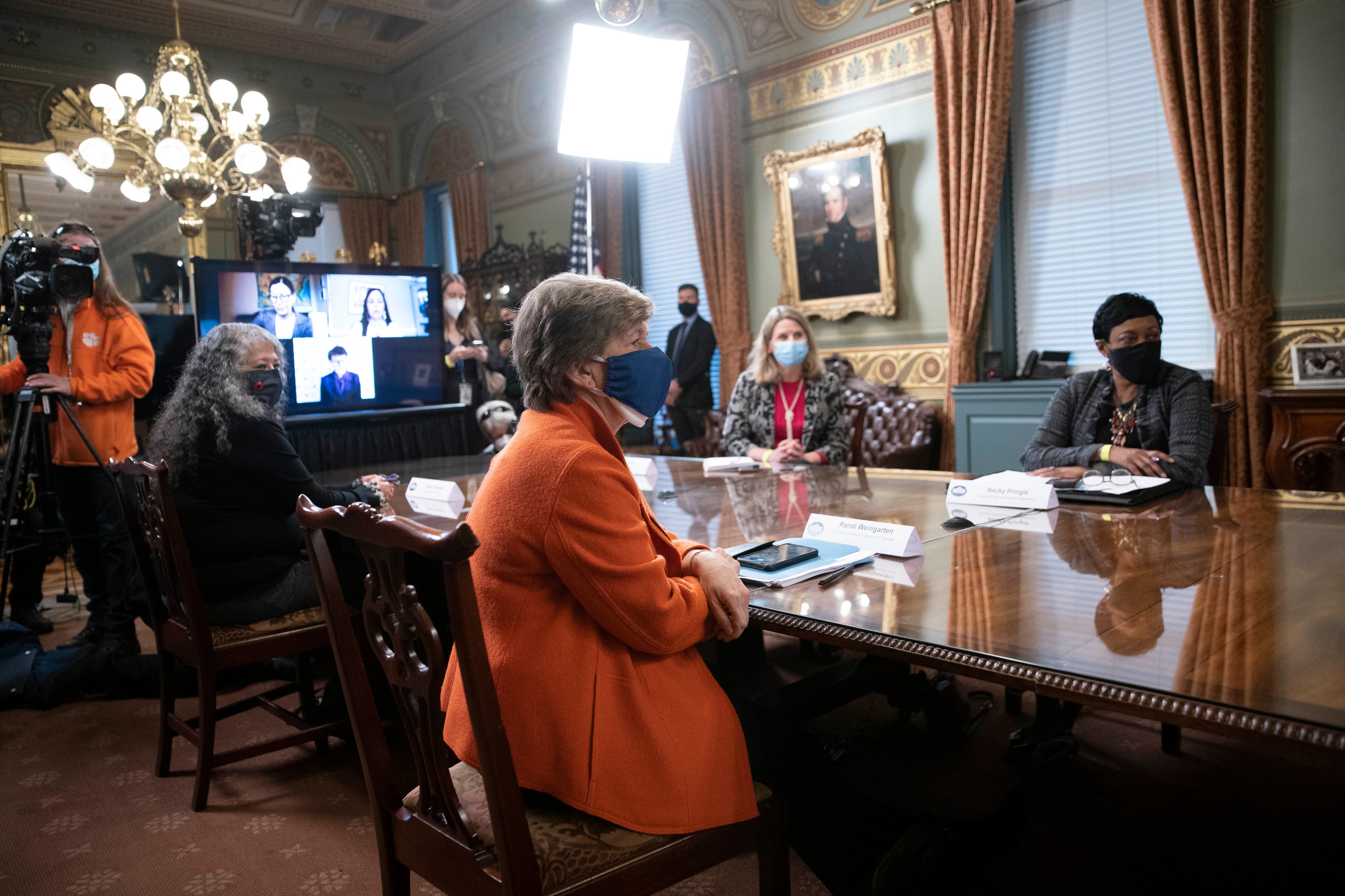 Four women sit around a table during a meeting to commemorate Women’s History Month. AFT President Randi Weingarten is on the left side of the table wearing an orange jacket and blue mask. There is a projection on a television in the background and a member of a film crew.