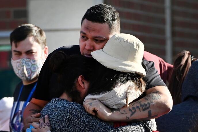 A large man with close-cropped hair tightly embraces two women who bury their heads on his shoulder. One woman has long dark hair pulled back and a gray sweater. The other is wearing a light tan bucket hat and jacket.