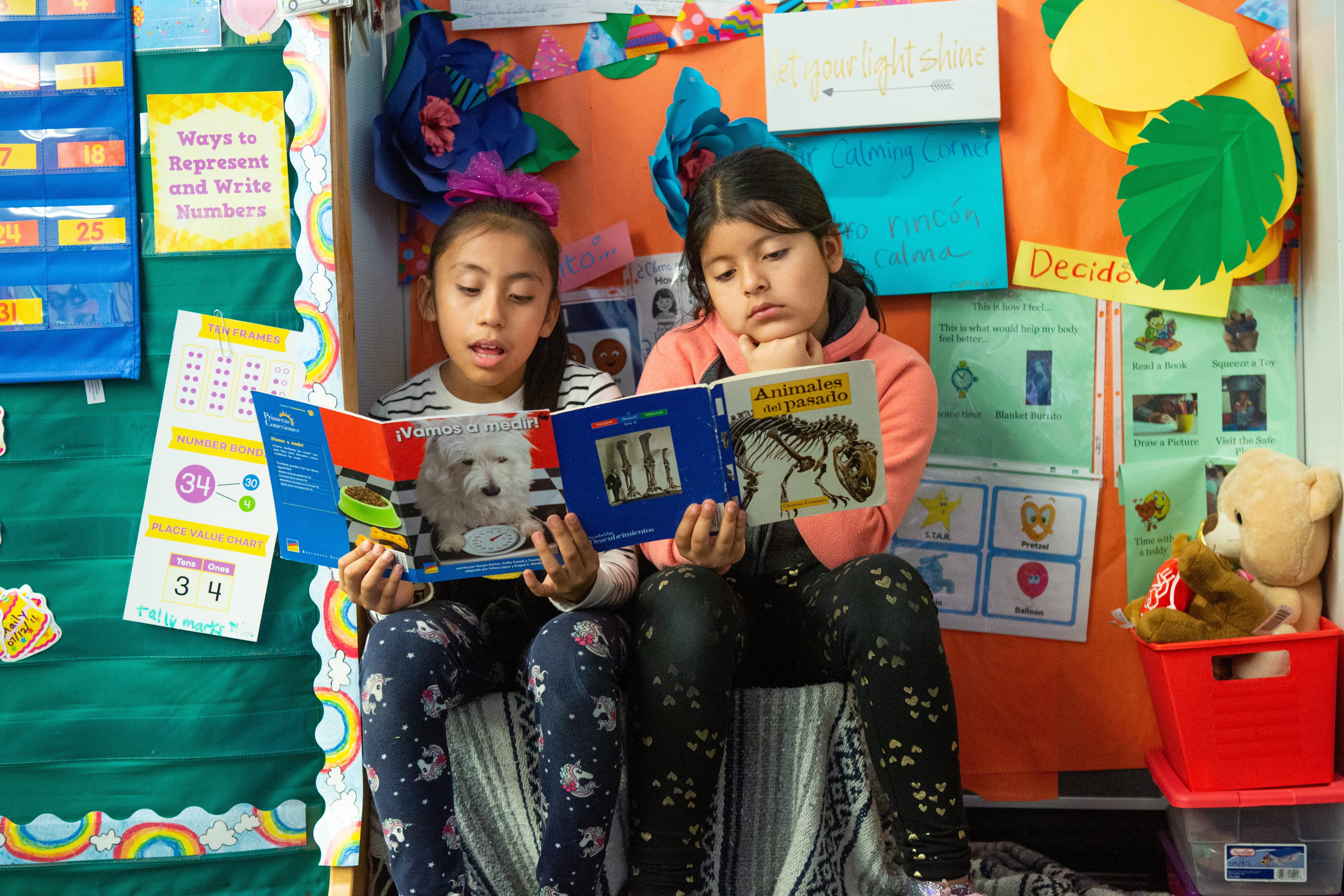 Two students sit next to each other reading a book with a colorful wall in the background.