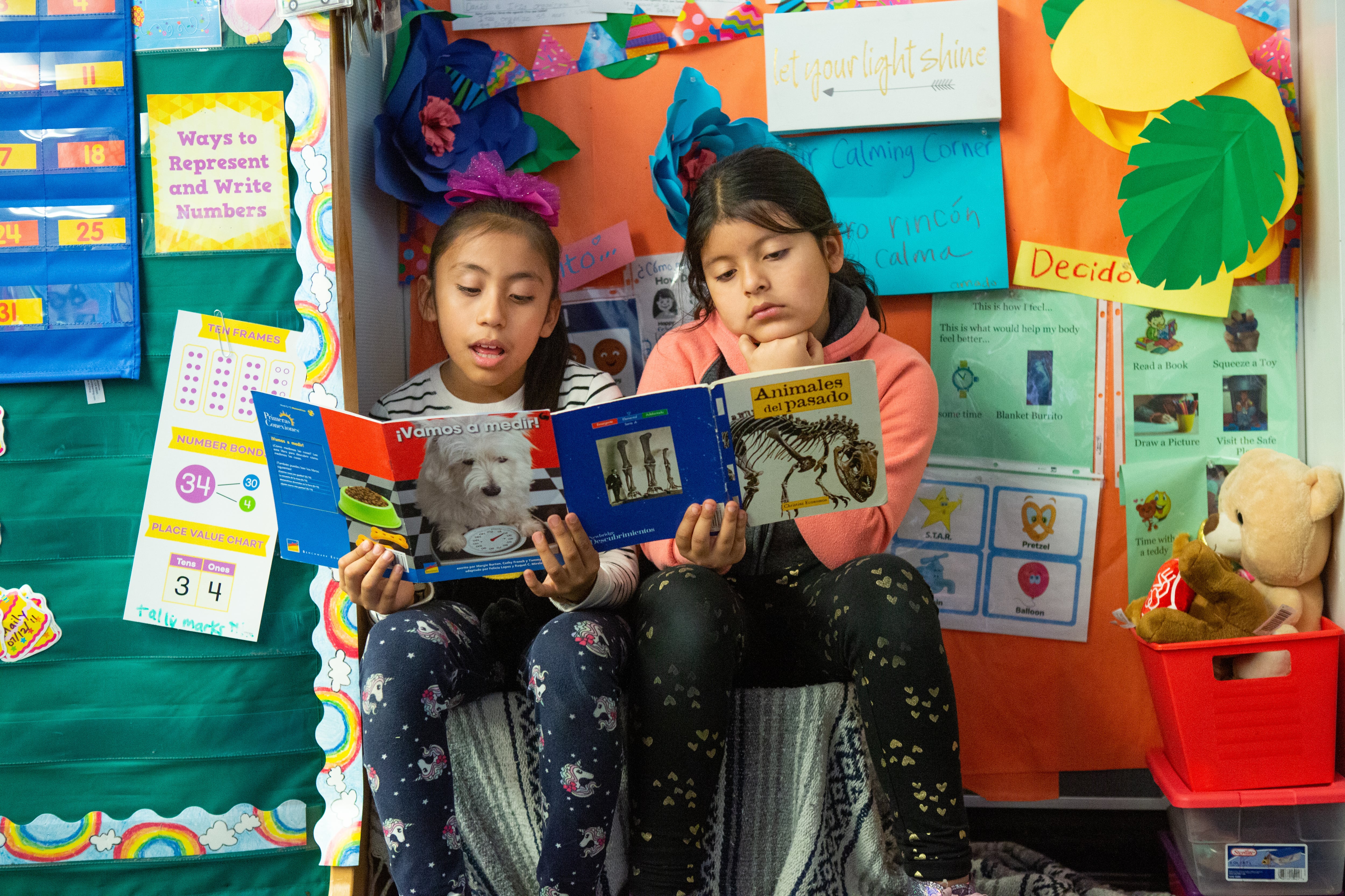 Two students sit next to each other reading a book with a colorful wall in the background.