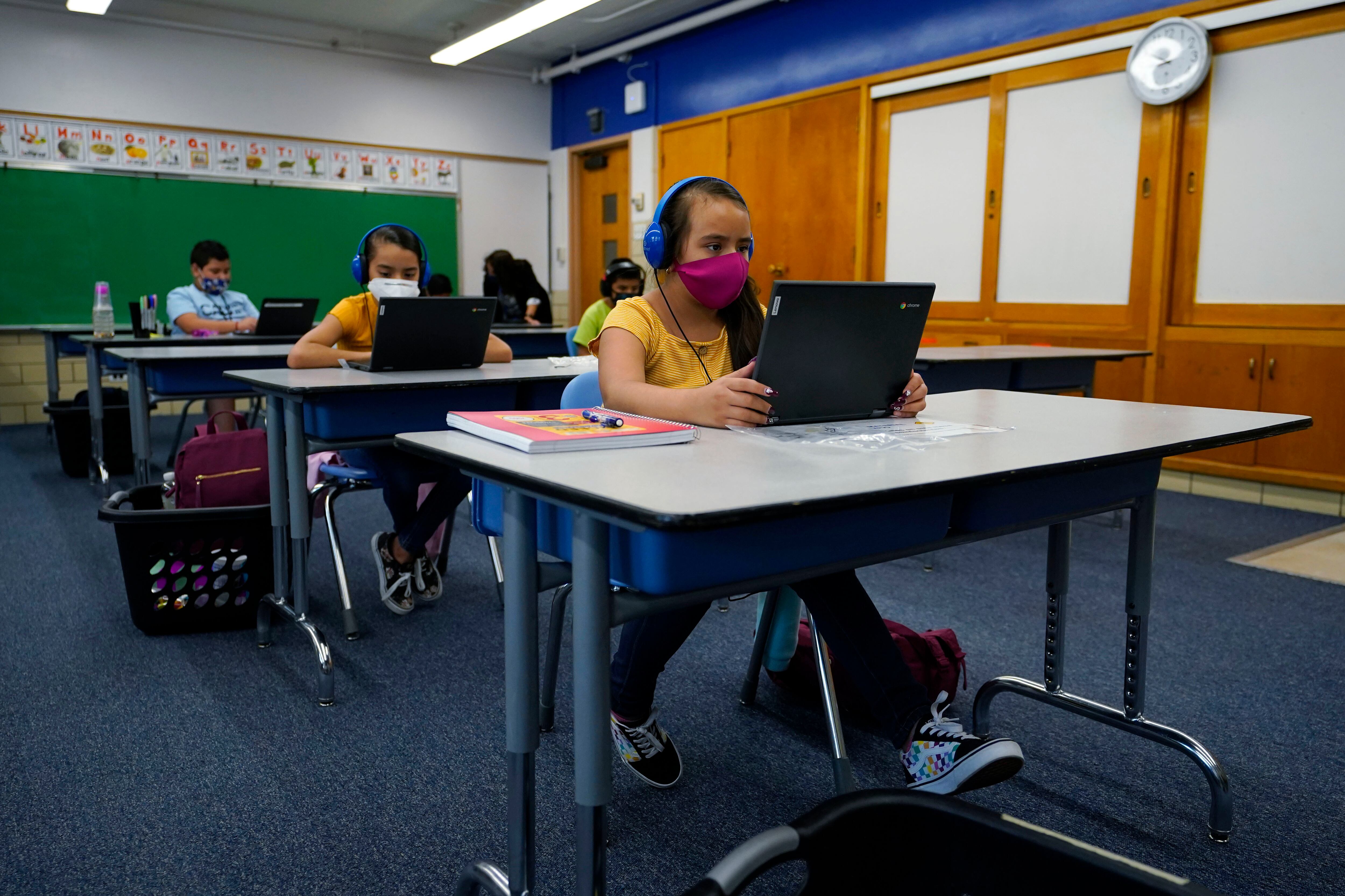 Masked students work on laptops at spaced out desks in a learning center.