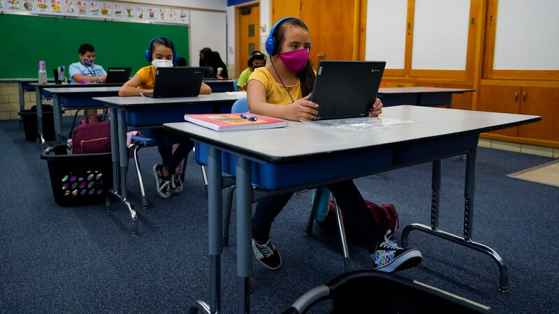 Masked students work on laptops at spaced out desks in a learning center.