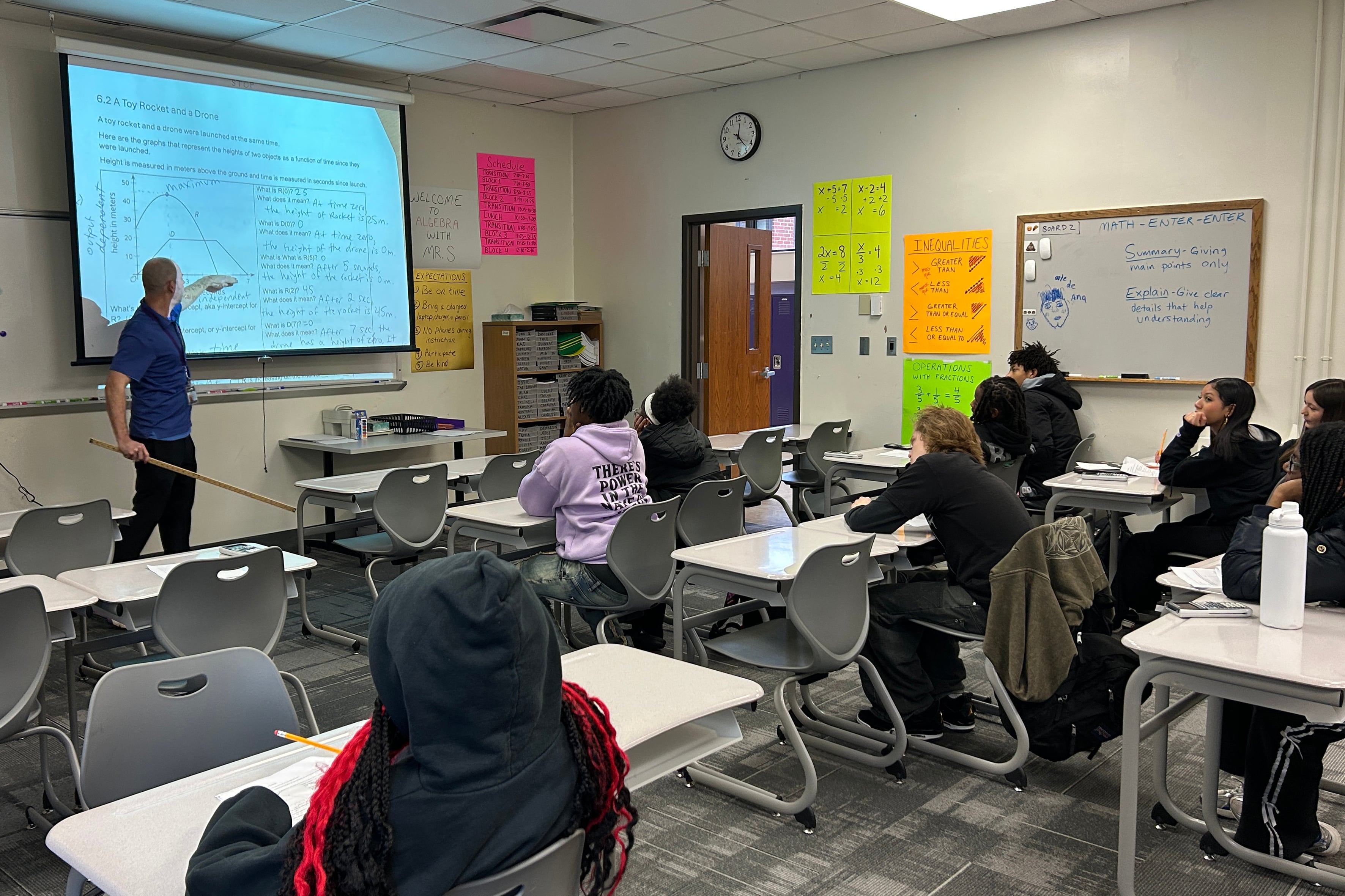 A photograph of a teacher pointing with a long ruler at a projector screen while a room full of high school students sit at their desks in a classroom.