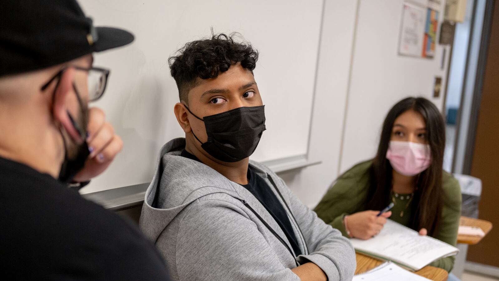 A young man, wearing a grey hoodie and black mask, speaks to his classmates at the desks on either side of him.