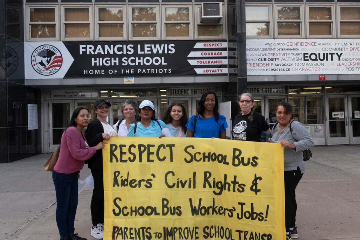 A group of people stand in front of a school holding a yellow sign.