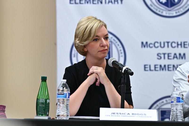 A white woman with short blonde hair sits at a table with a microphone in front of her.