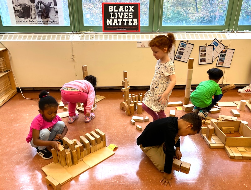 A girl in a beige dress stands in the middle of her classmates as they work on a brick activity.