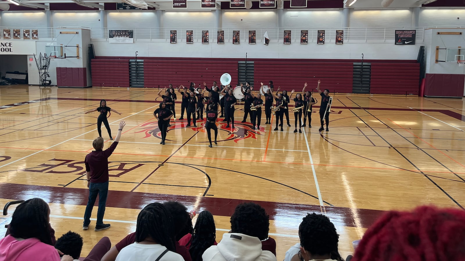 A marching band performs on the floor of the high school gym with students watching.