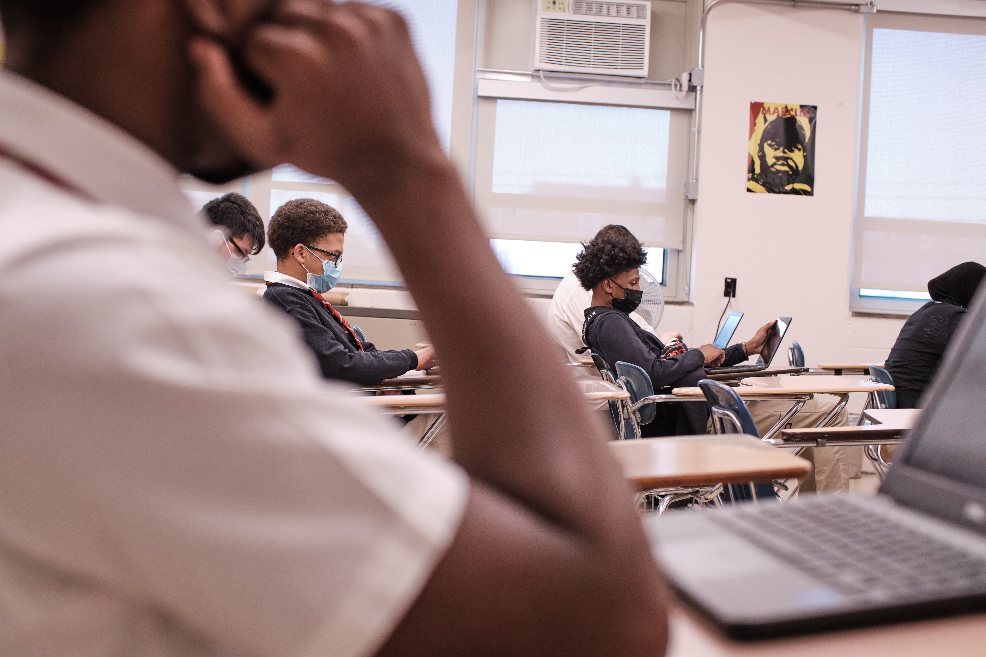 Students wearing masks sit at desks while looking at laptop computers.