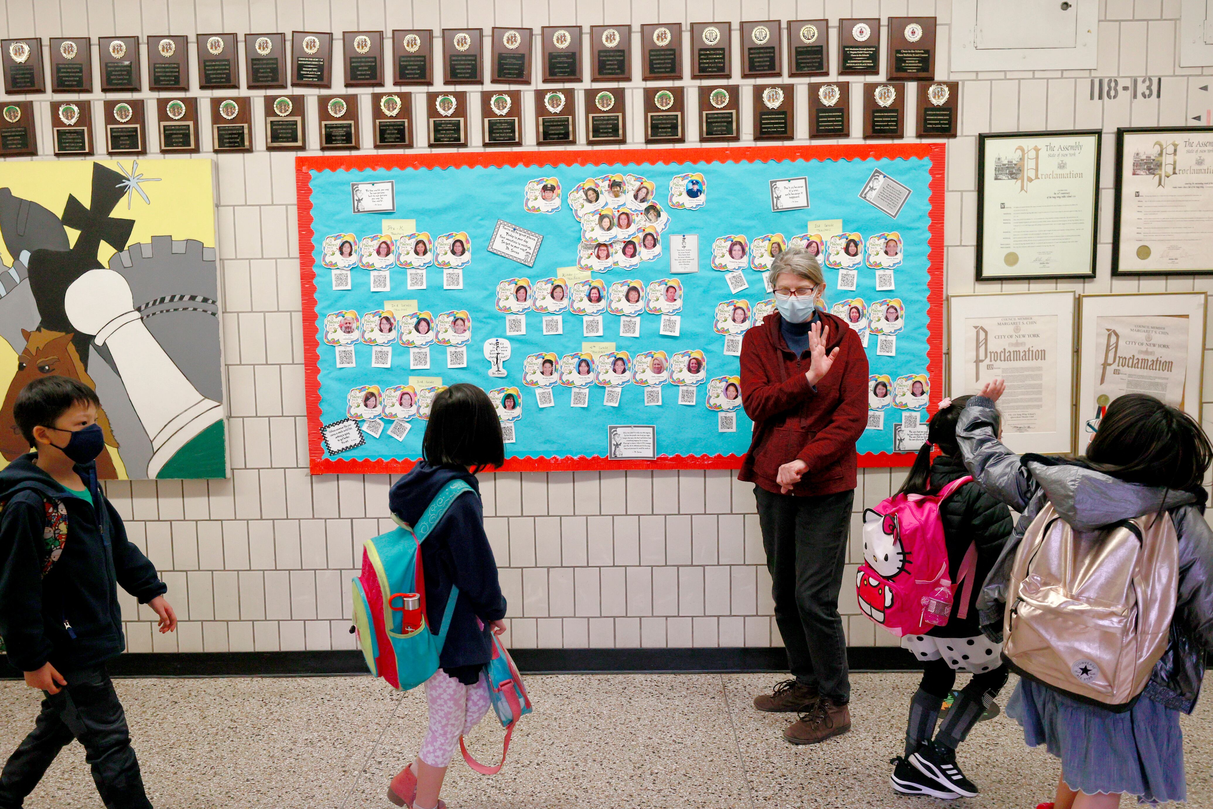 Four elementary school students with colorful backpacks walk by a middle-aged woman working as a paraprofessional and wave goodbye during school dismissal. The hallway is adorned with small awards and a sky blue board with student pictures on it.
