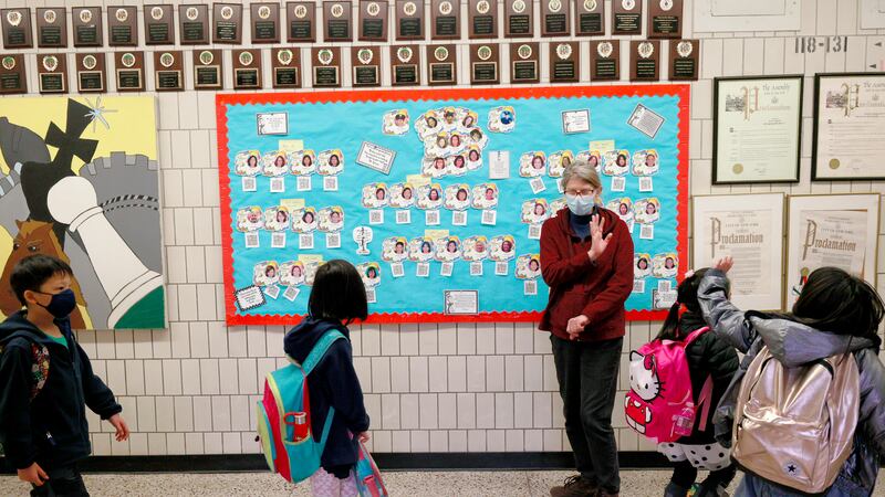 Four elementary school students with colorful backpacks walk by a middle-aged woman working as a paraprofessional and wave goodbye during school dismissal. The hallway is adorned with small awards and a sky blue board with student pictures on it.