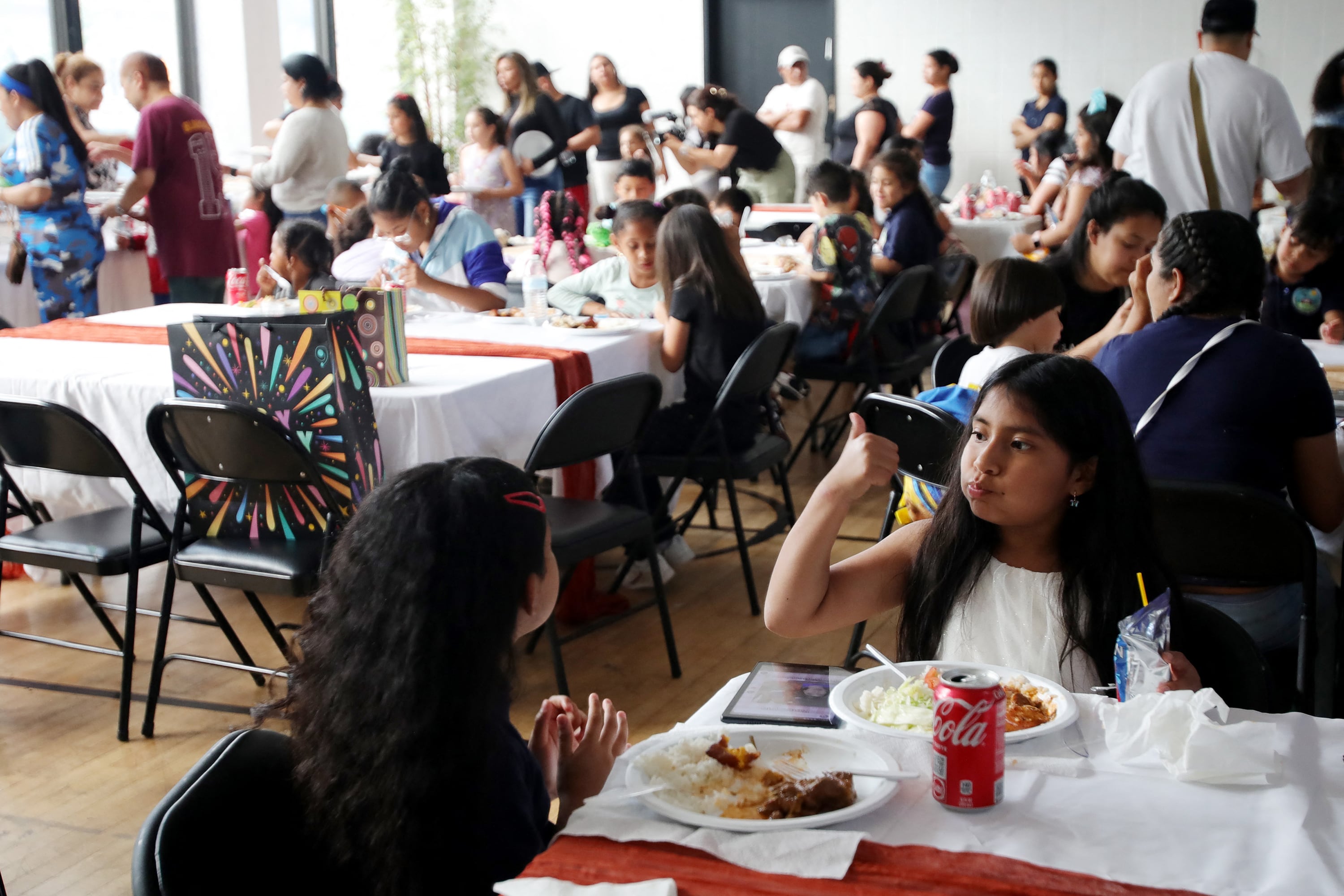Two young girls eat dinner at a table in the foreground while a large group of people, some standing and some sitting at tables in the background.