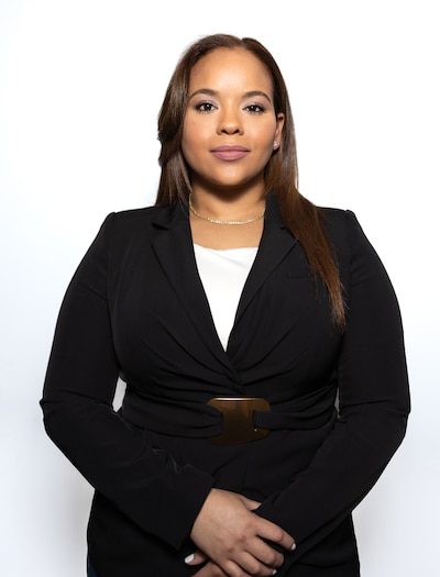A woman with long dark hair in a black suit poses for a portrait against a white background.