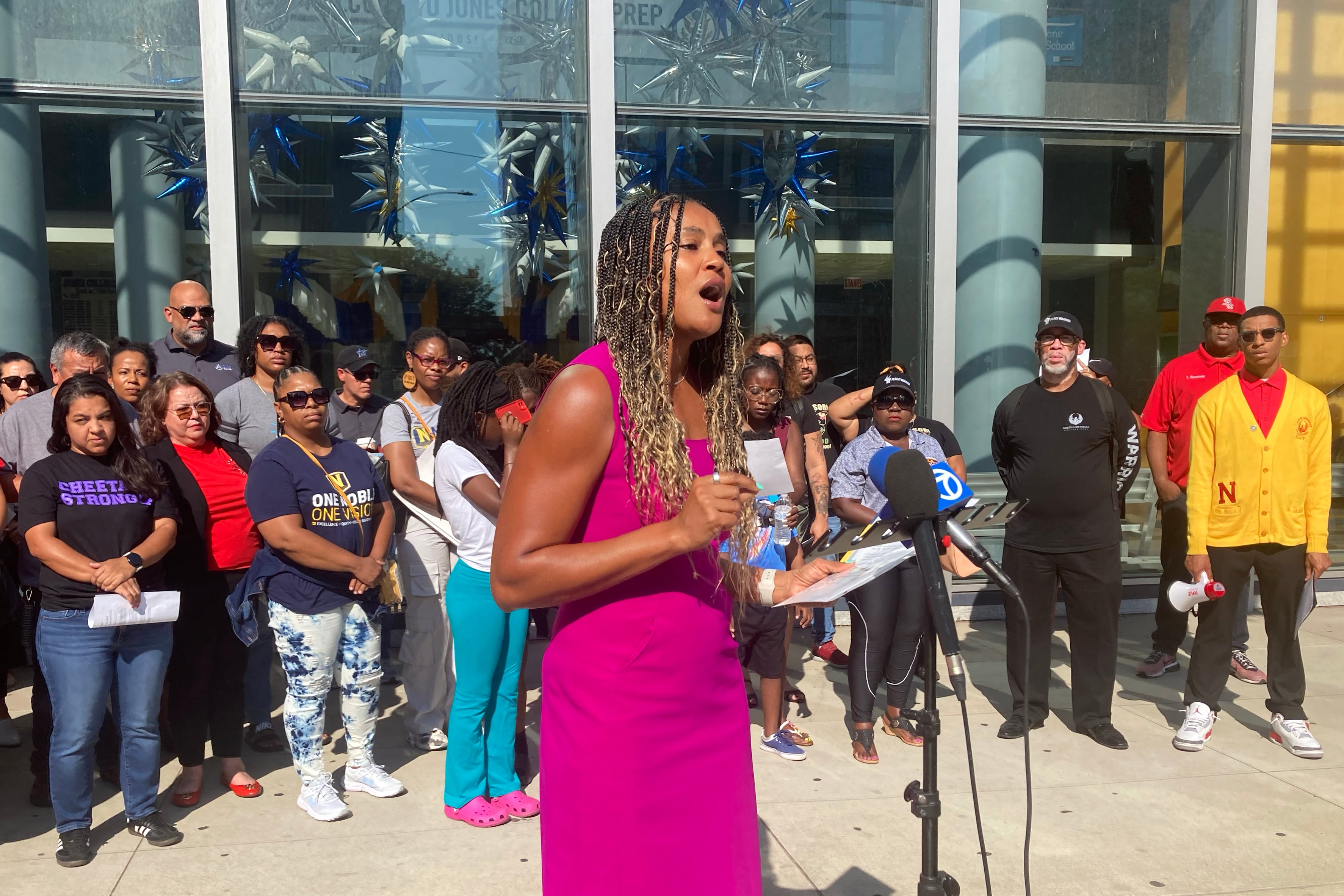 A woman with long braids and wearing a bright pink dress stands behind a microphone and in front of a large crowd of people outside.