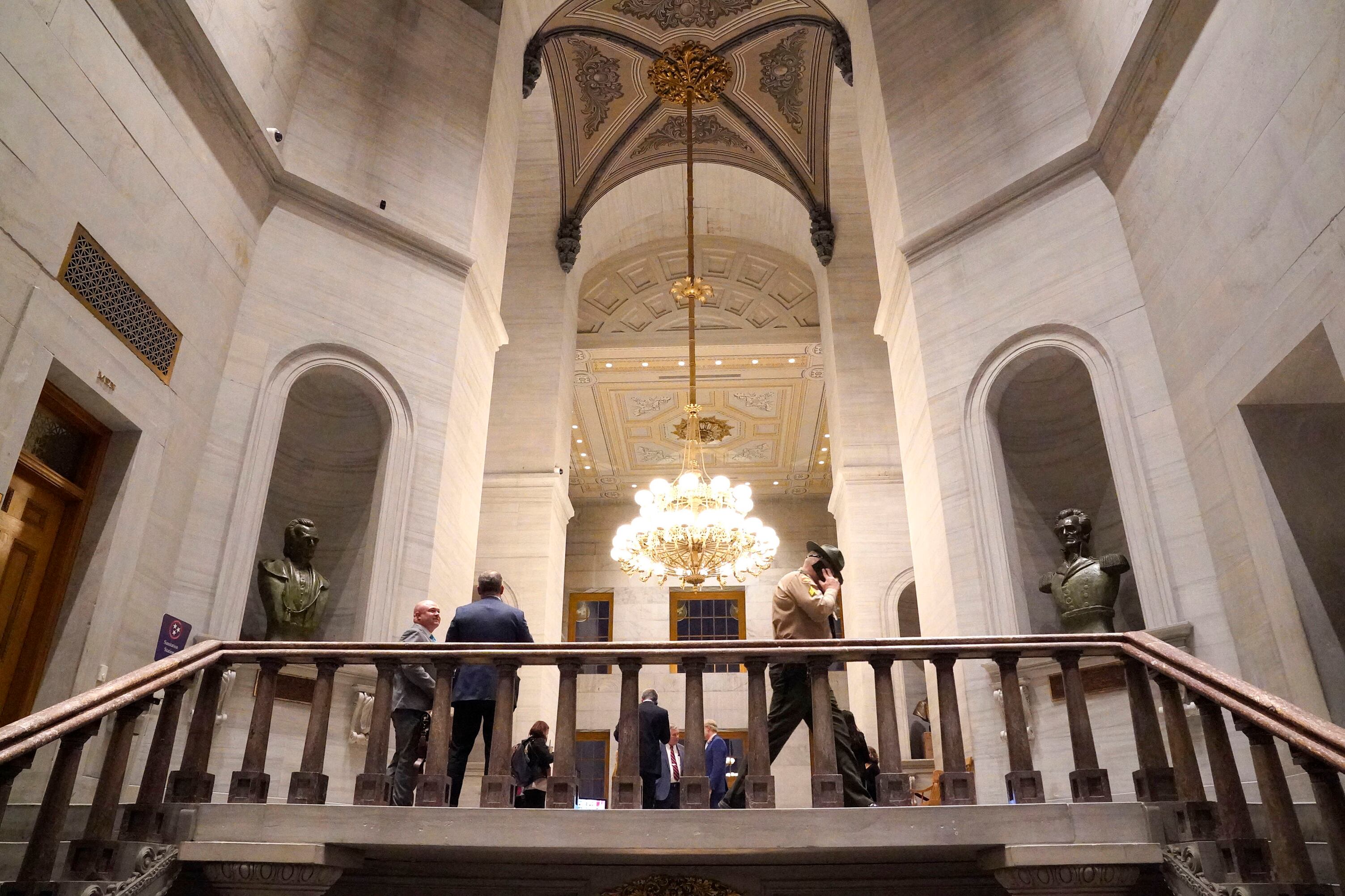 People mill inside of an ornate building lit by a large chandalier.