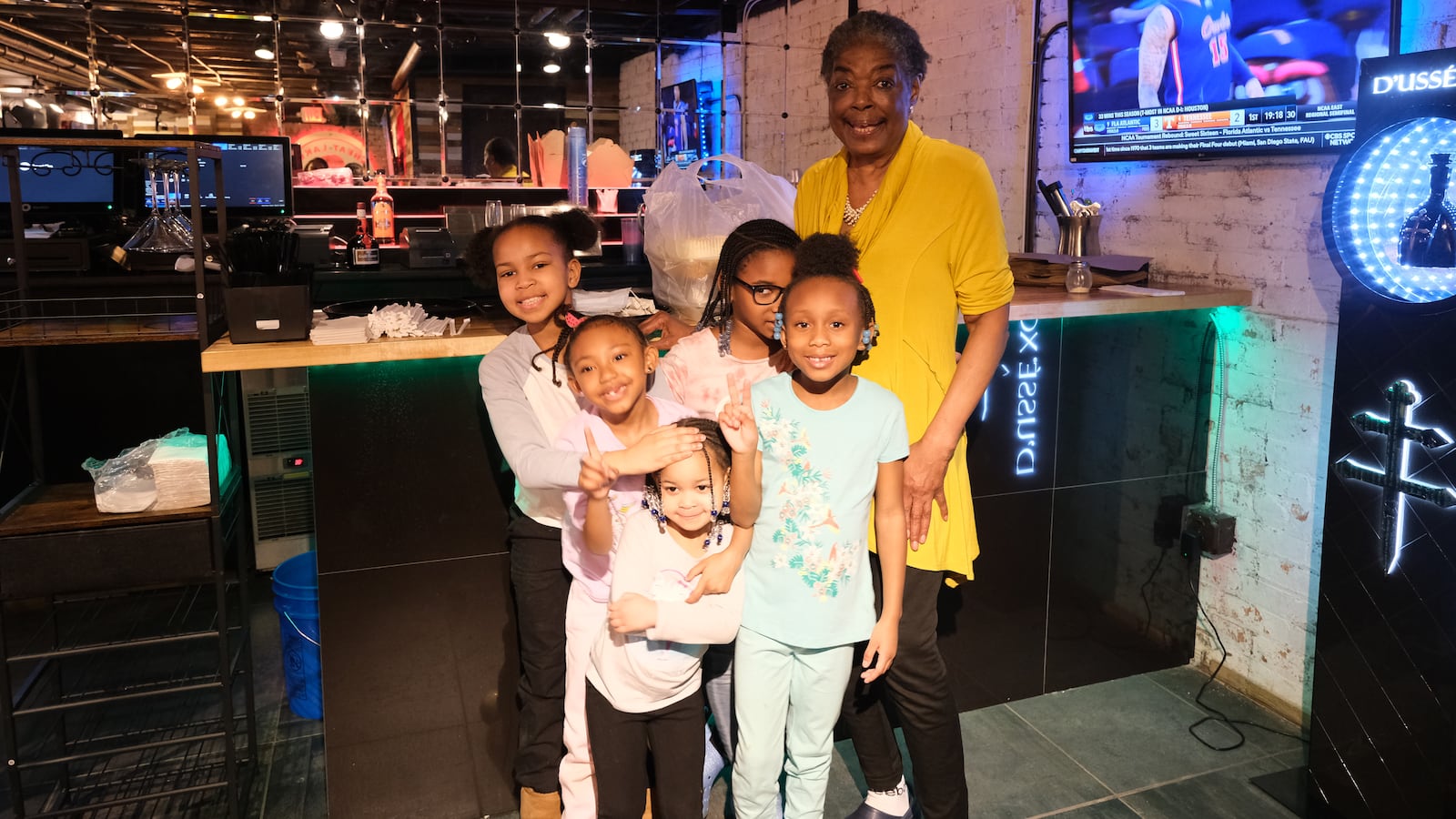 An older woman wearing a yellow shirt poses for a portrait with five young girls in the room of a restaurant with a tv on the wall.
