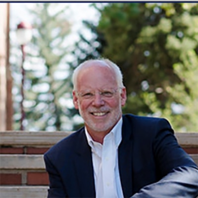 A photograph of a white man with glasses sitting outside.
